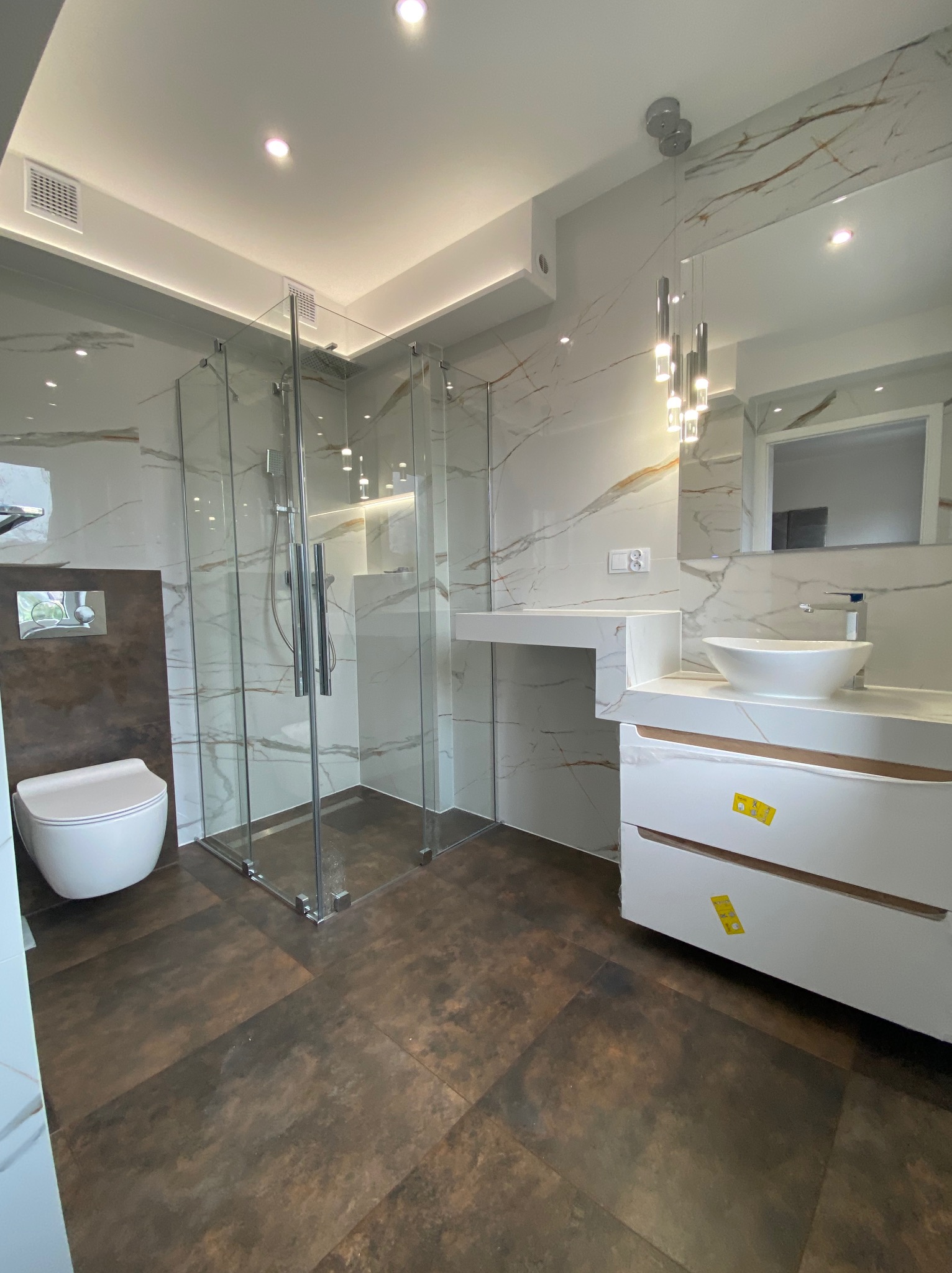 Nowoczesna łazienka z kabiną prysznicową typu walk-in, wiszącą toaletą, umywalką nablatową i brązowymi płytkami podłogowymi, widoczne żółte naklejki ochronne na szafce umywalkowej.