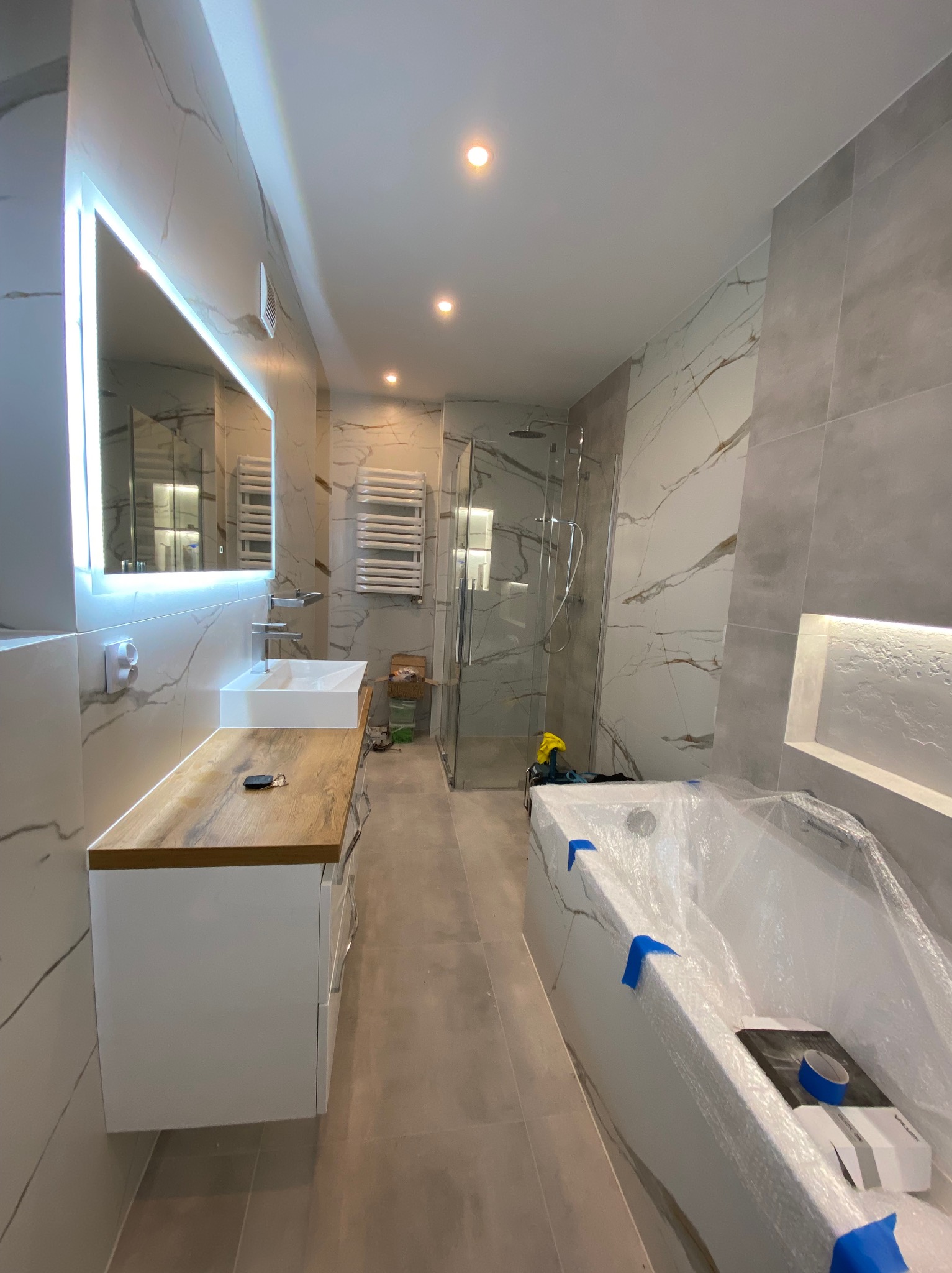 Wnętrze łazienki w trakcie prac wykończeniowych: obudowana wanna zabezpieczona folią, umywalka z drewnianym blatem i podświetlanym lustrem, płytki imitujące marmur na ścianach.