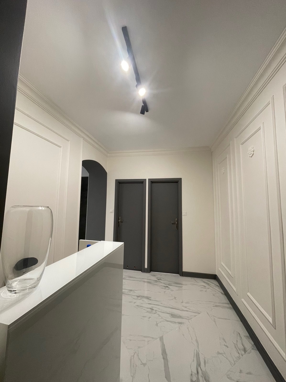 Nowoczesny korytarz z białymi panelami ściennymi, marmurową podłogą i ciemnymi drzwiami, oświetlony reflektorami sufitowymi.