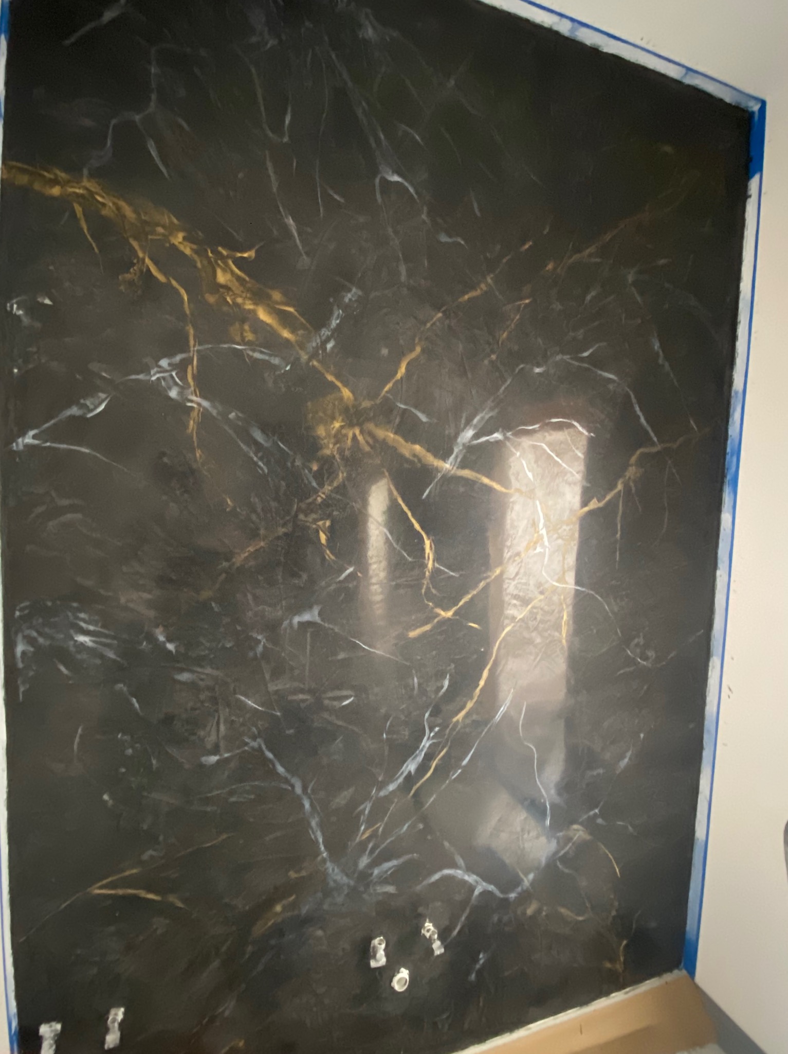 Ściana z czarną, błyszczącą fakturą imitującą marmur z białymi i złotymi żyłkami, zabezpieczona niebieską taśmą malarską na krawędziach.