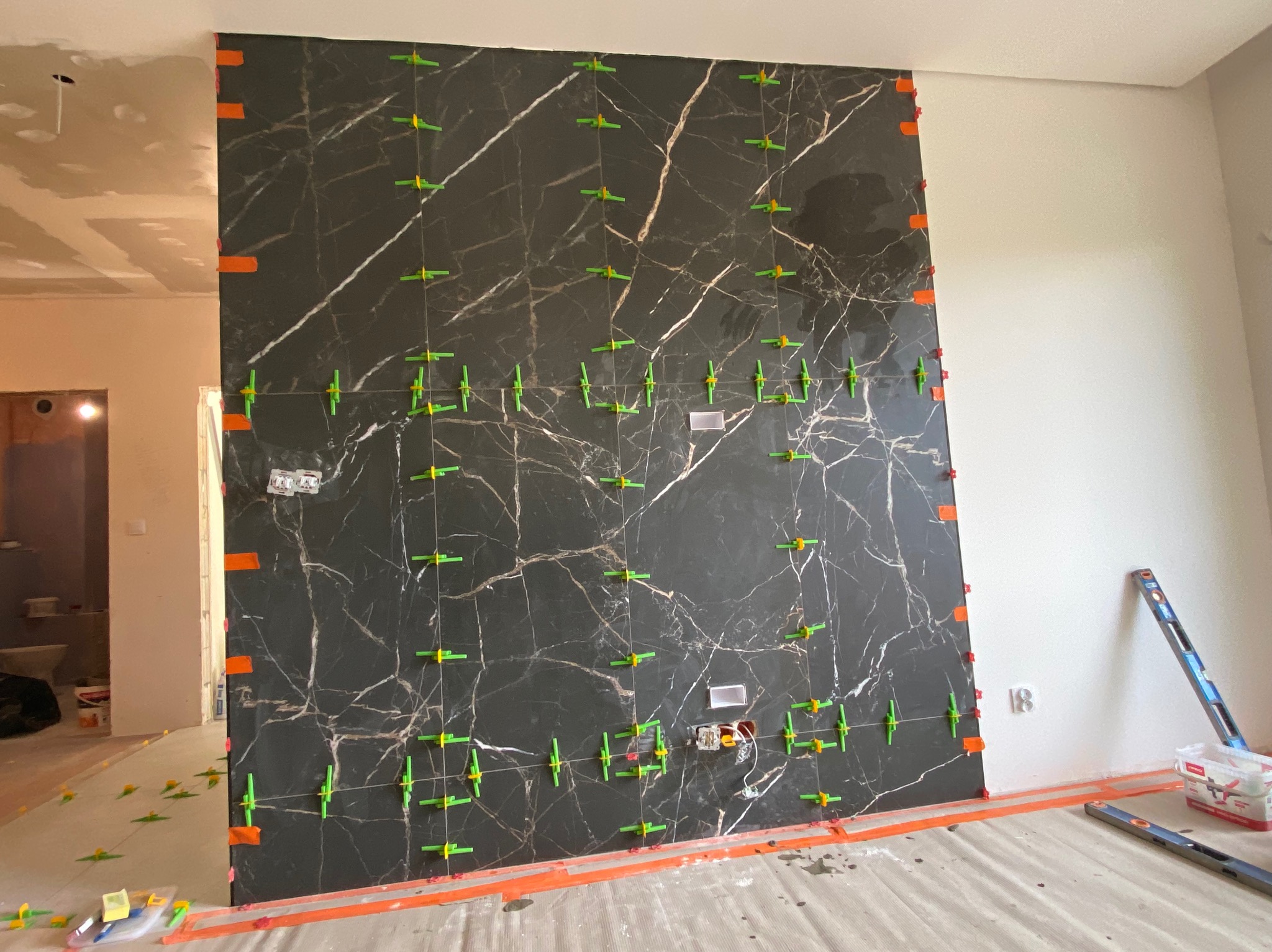 Ściana z czarnych płytek imitujących marmur, w trakcie montażu z użyciem zielonych krzyżyków dystansowych i pomarańczowych klinów poziomujących; widoczne przygotowane punkty elektryczne.