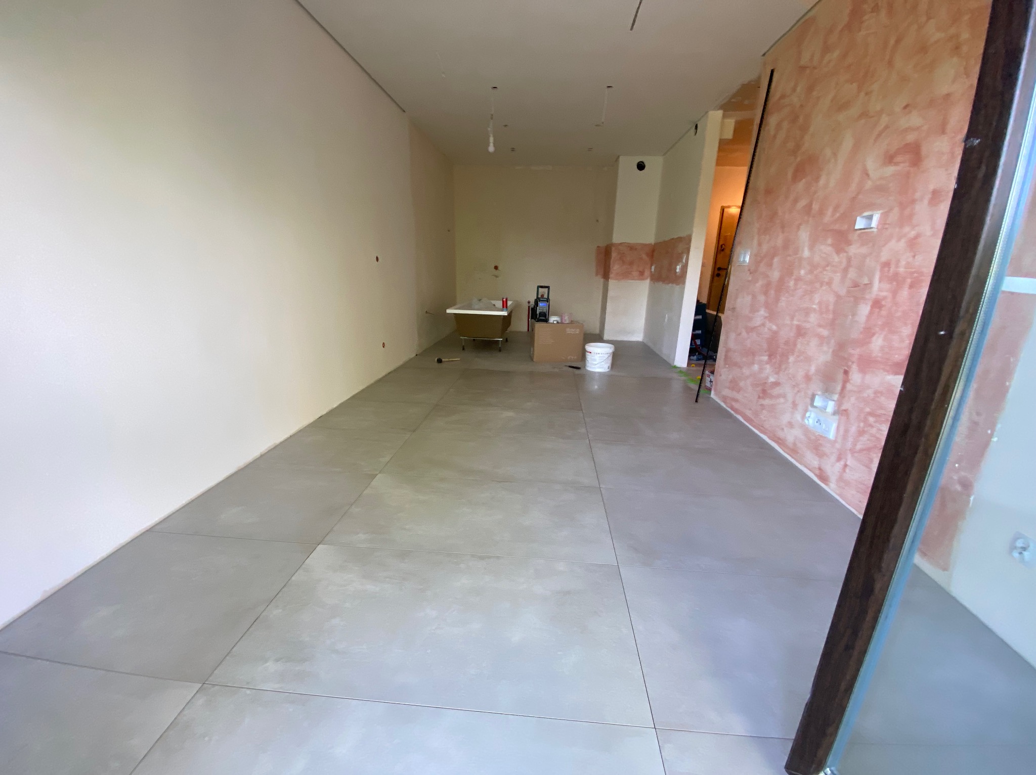 Przestronne wnętrze w trakcie prac wykończeniowych: ułożone szare płytki podłogowe, ściany częściowo pomalowane na beżowo i różowo, wolnostojąca wanna na nóżkach w oddali, widoczne materiały...