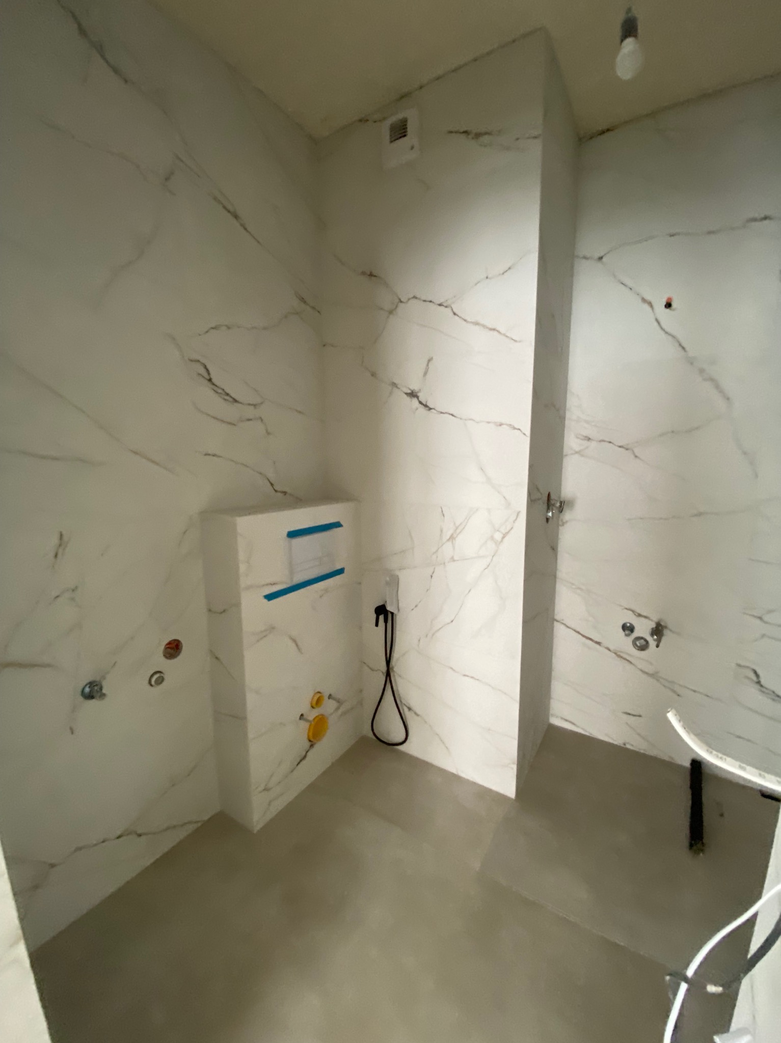 Wnętrze łazienki w trakcie prac wykończeniowych w Katowicach, widoczne białe marmurowe płytki na ścianach, zabudowa stelaża WC z niebieską taśmą, przygotowane instalacje wodno-kanalizacyjne...