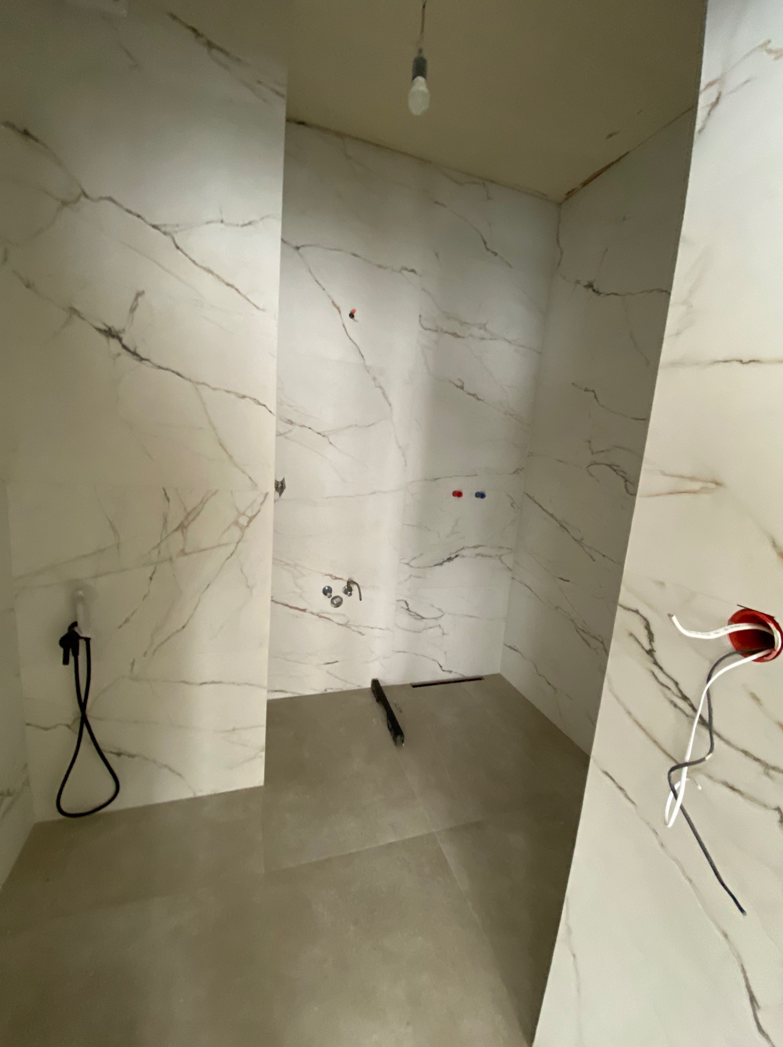 Wnętrze łazienki w trakcie prac wykończeniowych: białe płytki imitujące marmur na ścianach, szara podłoga, widoczne instalacje wodne i elektryczne, surowa żarówka zwisająca z sufitu.