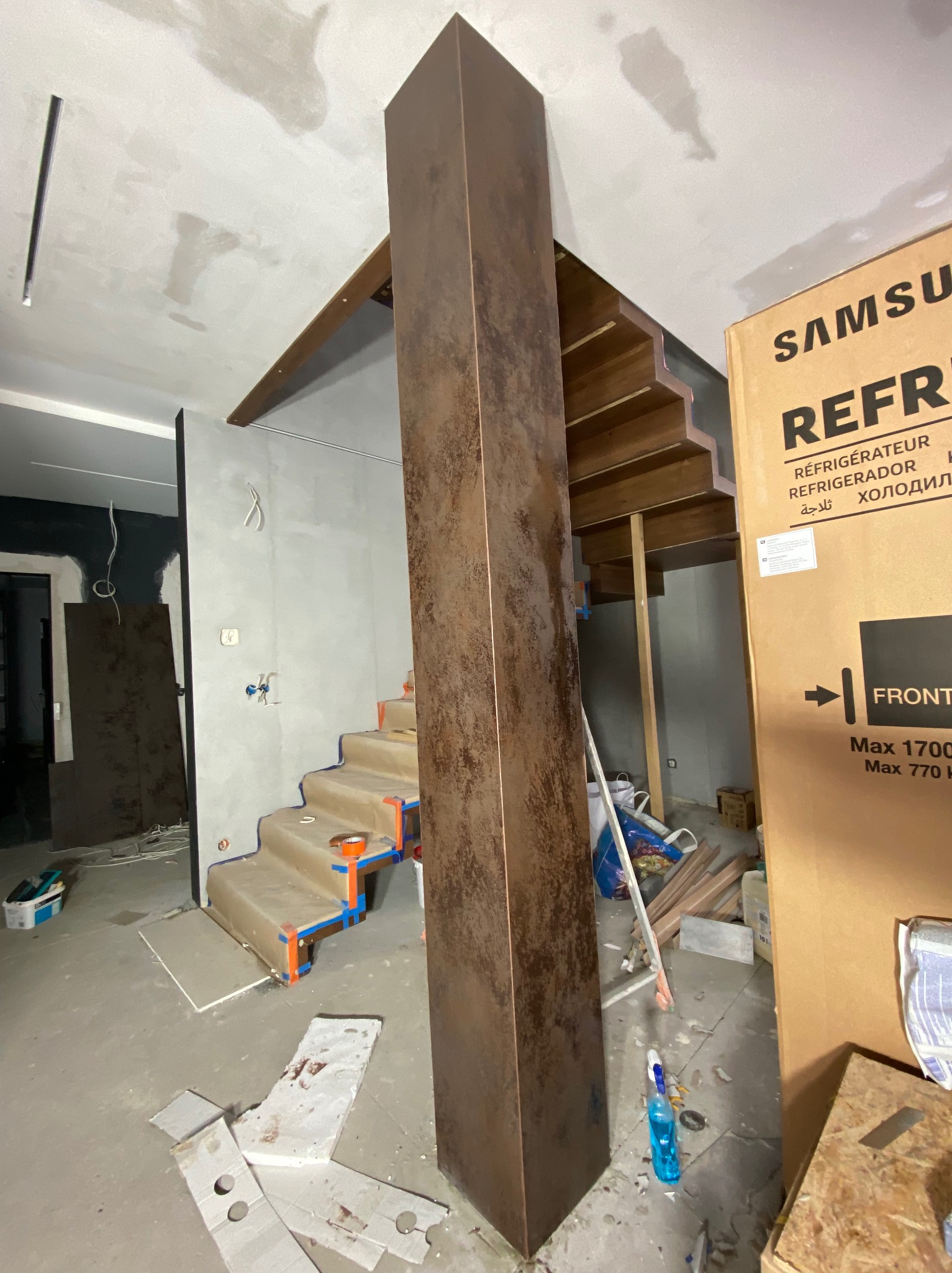 Wnętrze w trakcie prac wykończeniowych: zabezpieczone schody z drewnianymi stopniami, pionowa kolumna o metalicznym wykończeniu, surowy sufit z widocznymi śladami po remoncie, karton z lodówki...