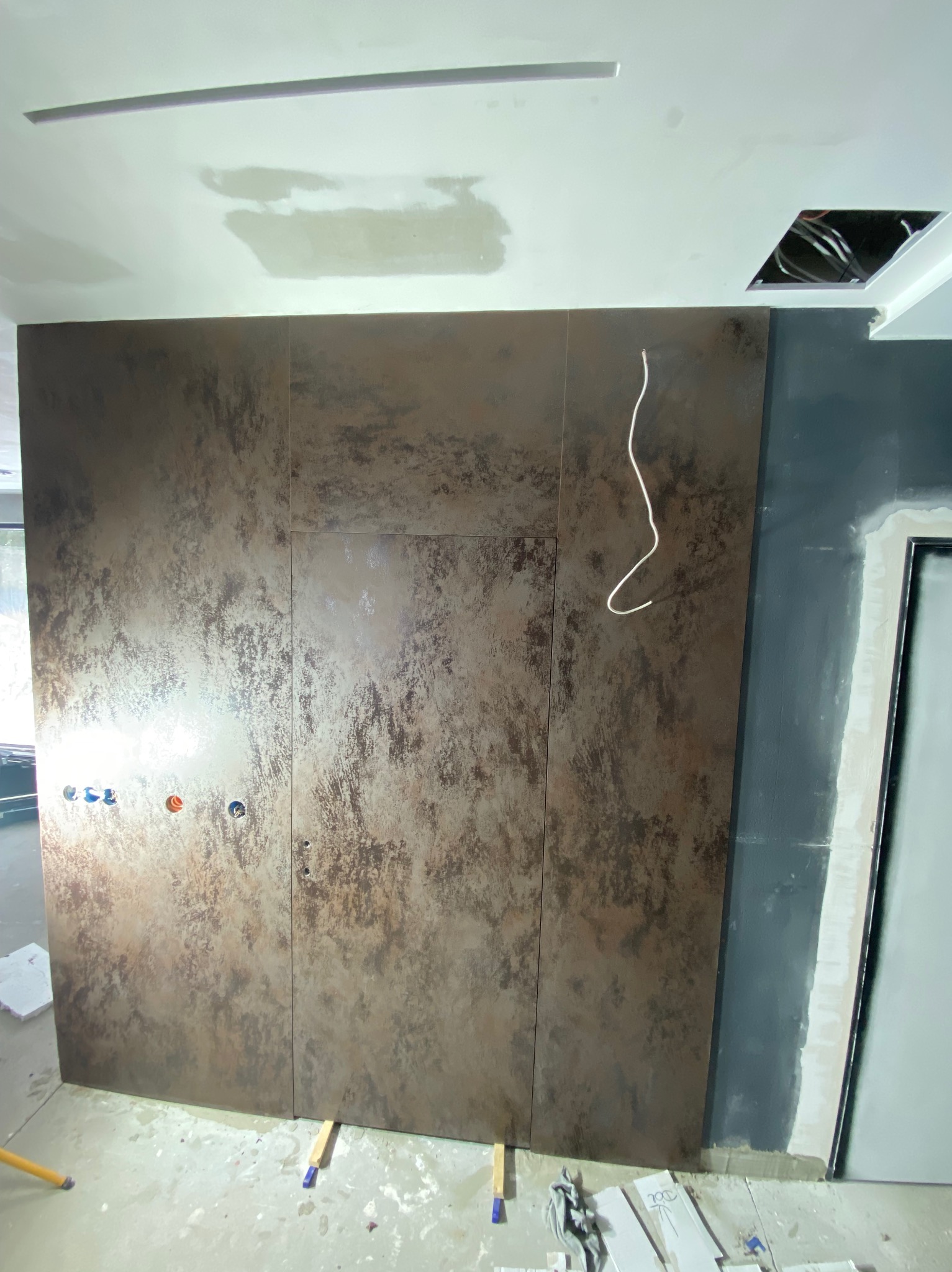 Montaż dużych, brązowo-beżowych płyt imitujących rdzę na ścianie w trakcie prac wykończeniowych. Widoczne otwory na instalacje, przewody elektryczne oraz fragment sufitu z niezamaskowanymi kablami.