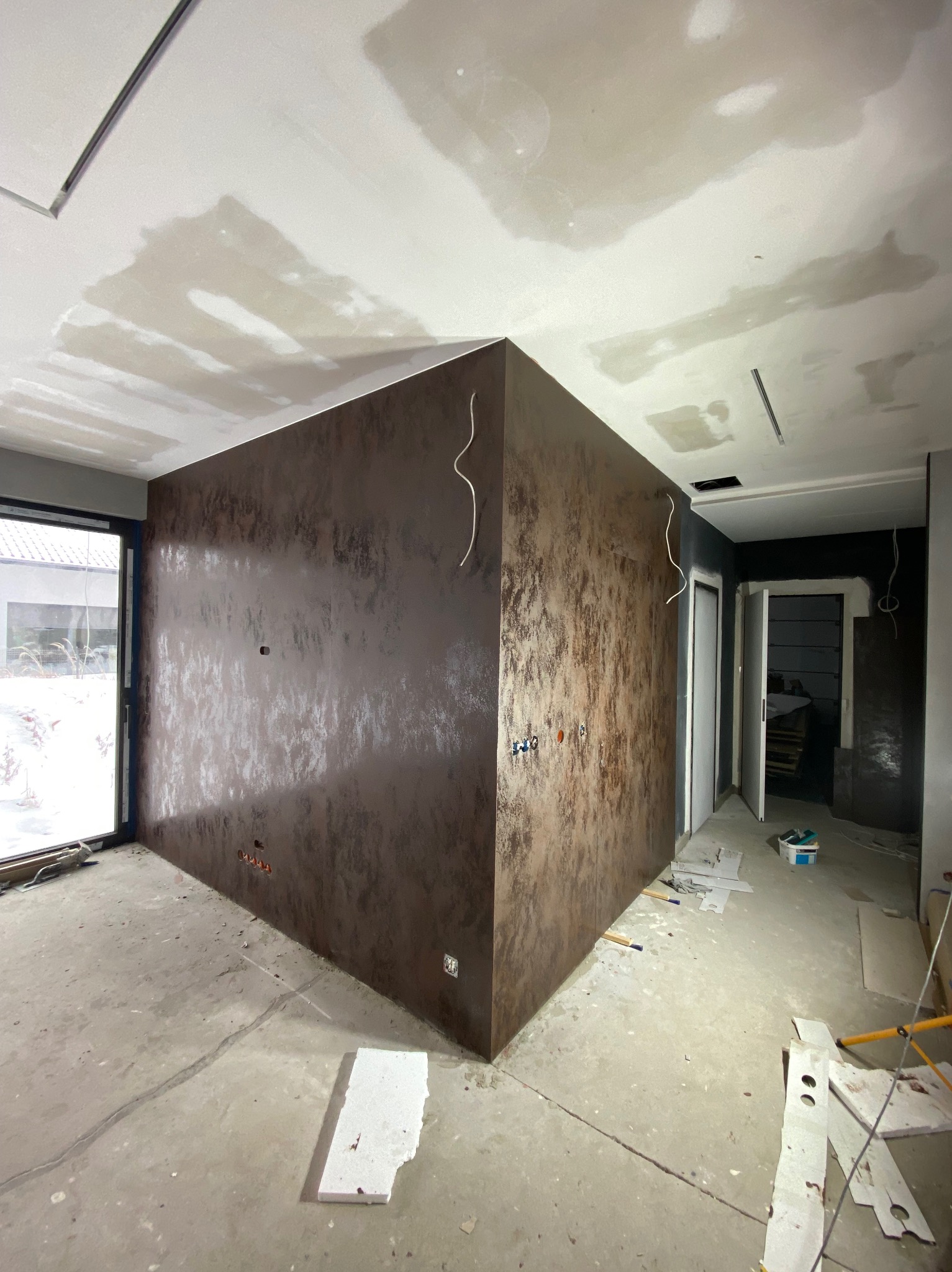 Pokój w trakcie prac wykończeniowych: ściany pokryte dekoracyjnym tynkiem w odcieniach brązu i beżu, sufit z widocznymi śladami szpachlowania, widoczne kable elektryczne, podłoga surowa, okno...