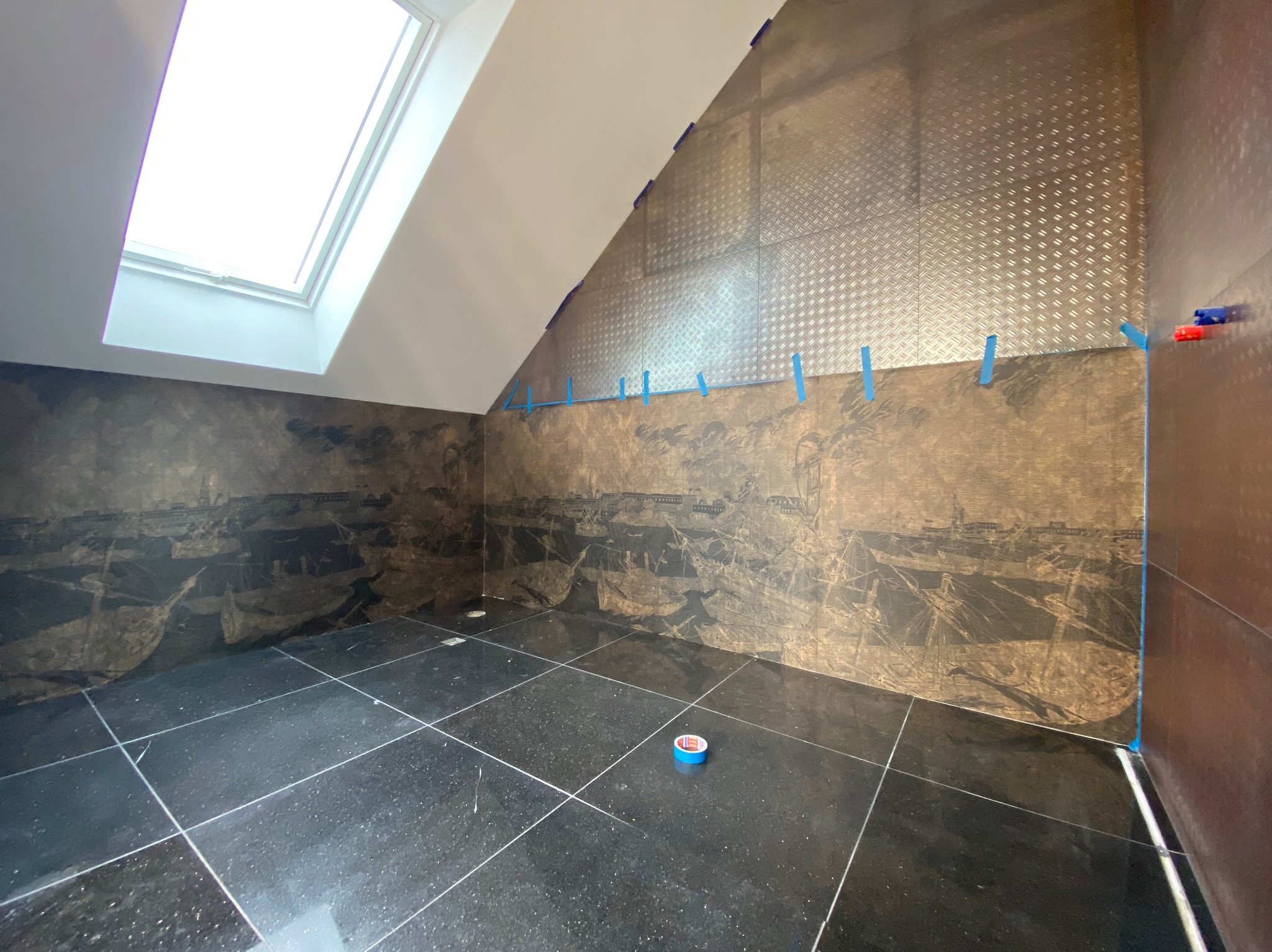 Łazienka w trakcie prac: czarne płytki podłogowe z białymi fugami, ściany częściowo wyłożone płytkami z motywem graficznym zabezpieczone taśmą malarską, poddasze z oknem dachowym, fragment ściany...