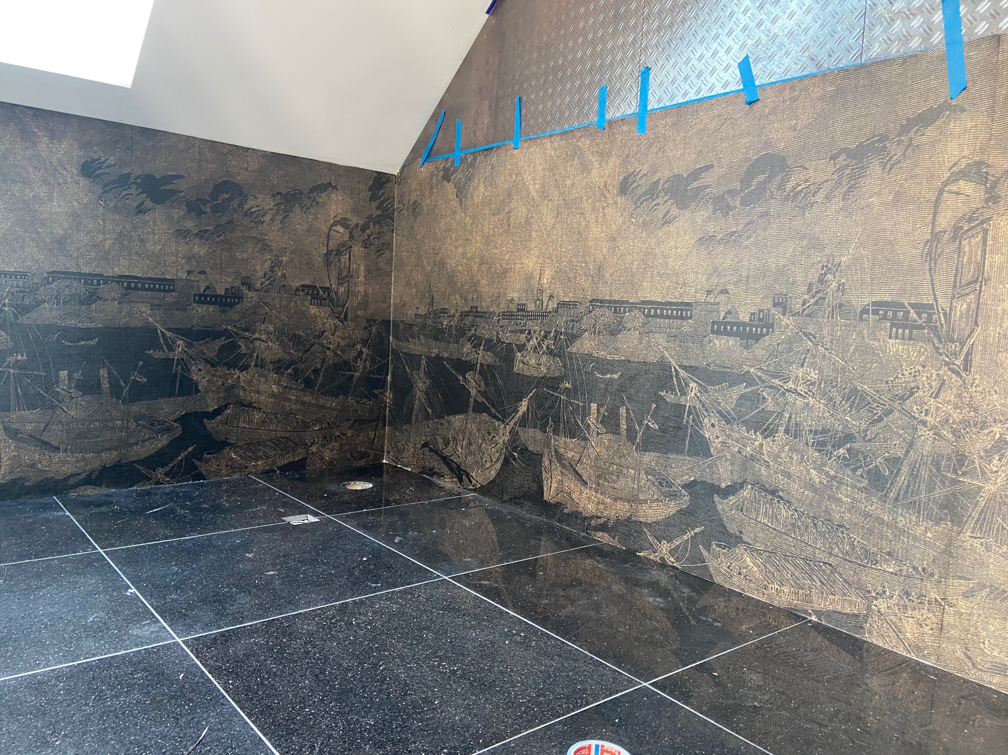 Ściana łazienki z tapetą w stylu marynistycznym przedstawiającą żaglowce i miasto portowe, połączona z górną częścią ściany wykończoną metalową blachą ryflowaną, zabezpieczoną niebieską taśmą...