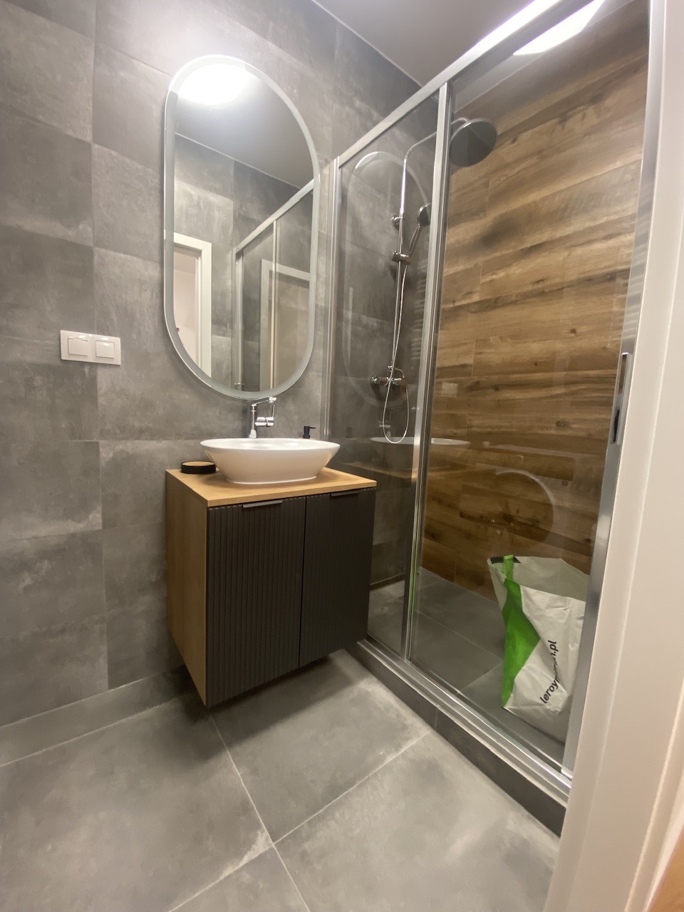 Nowoczesna łazienka z szarą płytką, owalnym lustrem z podświetleniem LED, umywalką nablatową na szafce z ciemnymi lamelami i kabiną prysznicową z drewnopodobną okładziną, torba zakupowa w kabinie.