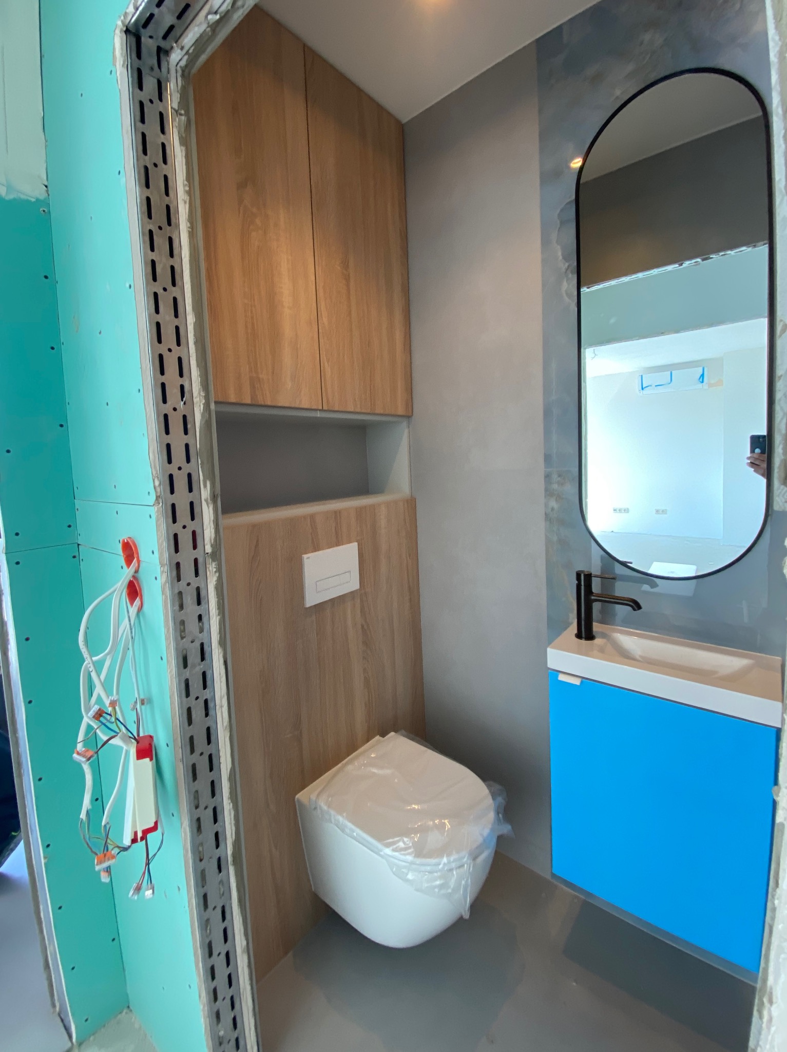 Wnętrze łazienki w trakcie prac: zabudowa WC z drewnianym wykończeniem, niebieska szafka pod umywalką, lustro w czarnej ramie. Na ścianie widoczne przewody elektryczne.