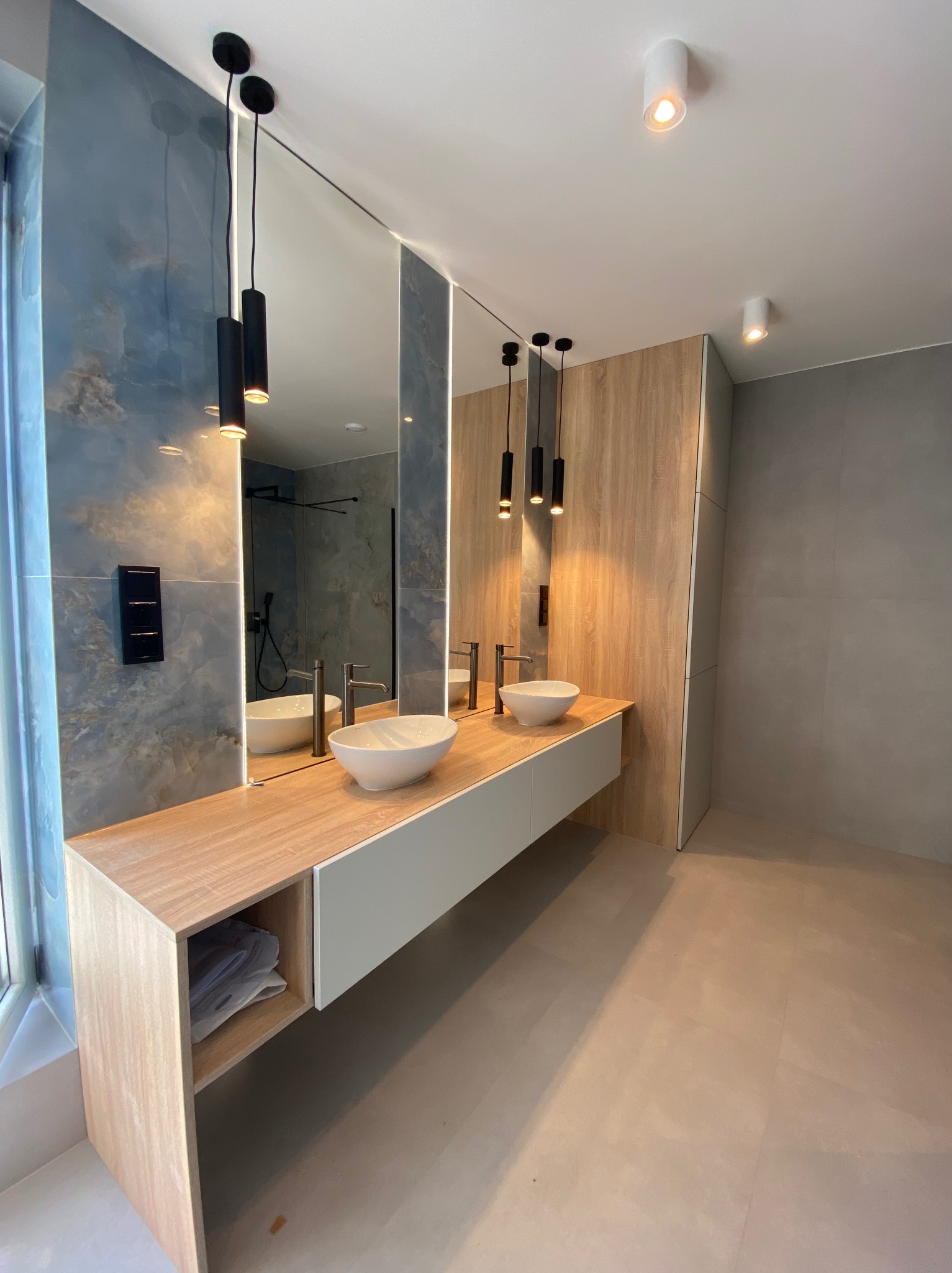 Nowoczesna łazienka z dwiema umywalkami nablatowymi, drewnianym blatem i dekoracyjną ścianą w odcieniach błękitu. Minimalistyczne oświetlenie i duże lustra.