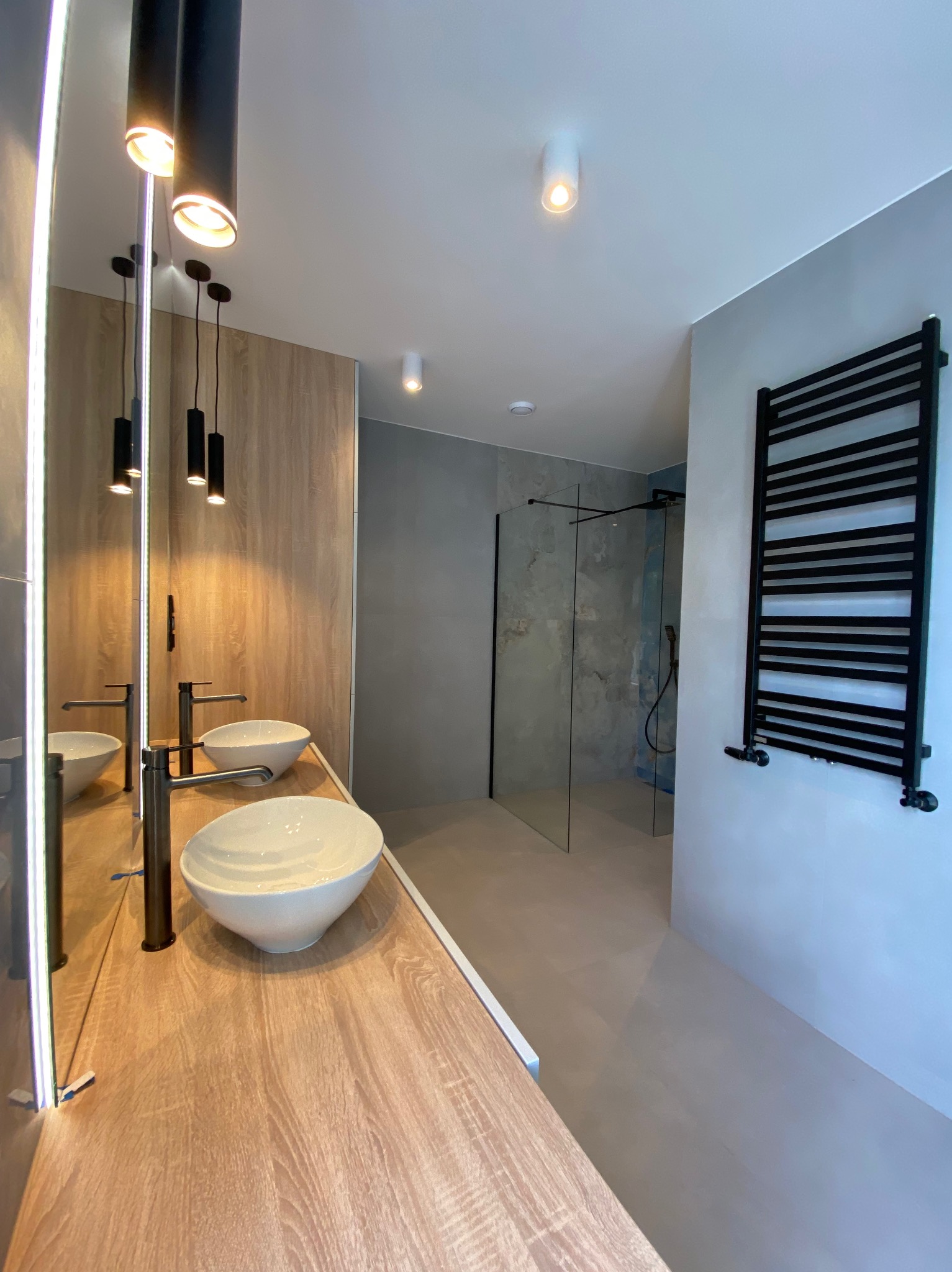 Nowoczesna łazienka z umywalkami nablatowymi, drewnianym blatem, szklaną kabiną prysznicową i czarnym grzejnikiem drabinkowym na szarej ścianie.