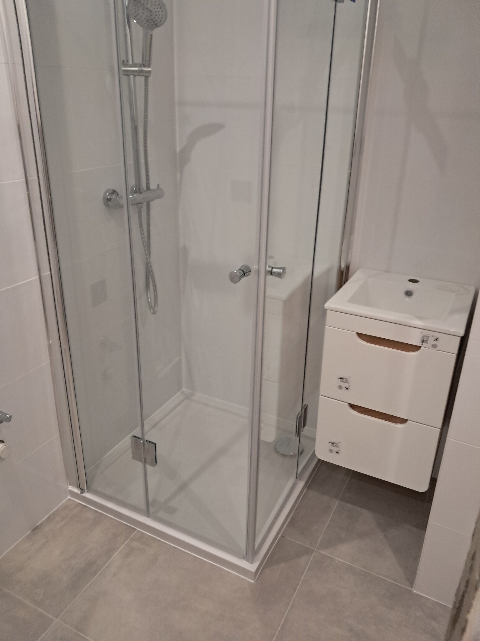 Nowoczesna kabina prysznicowa z białym brodzikiem i szklanymi drzwiami, obok biała szafka pod umywalkę z dwiema szufladami, szare płytki podłogowe.