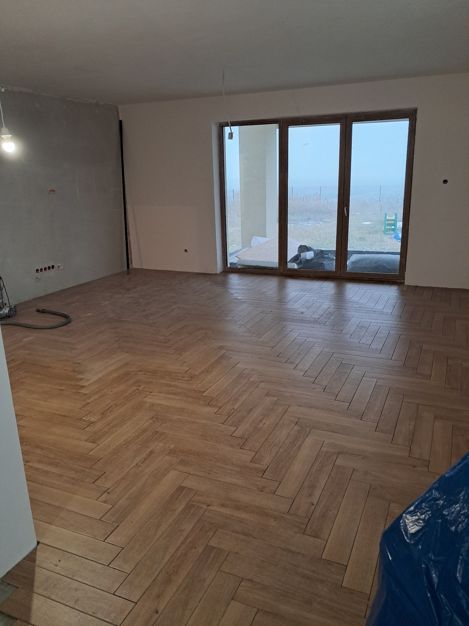 Ułożona podłoga z paneli w jodełkę w nowym, jasnym pomieszczeniu z dużym oknem tarasowym; widoczne ślady prac wykończeniowych na ścianach i sprzęt budowlany w rogu.