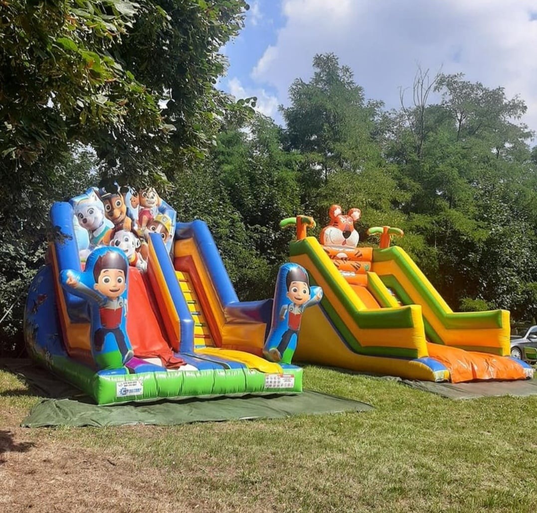 Dwa kolorowe dmuchane zamki ze zjeżdżalniami na trawie, jeden z motywem Psiego Patrolu, drugi z tygrysem na szczycie.