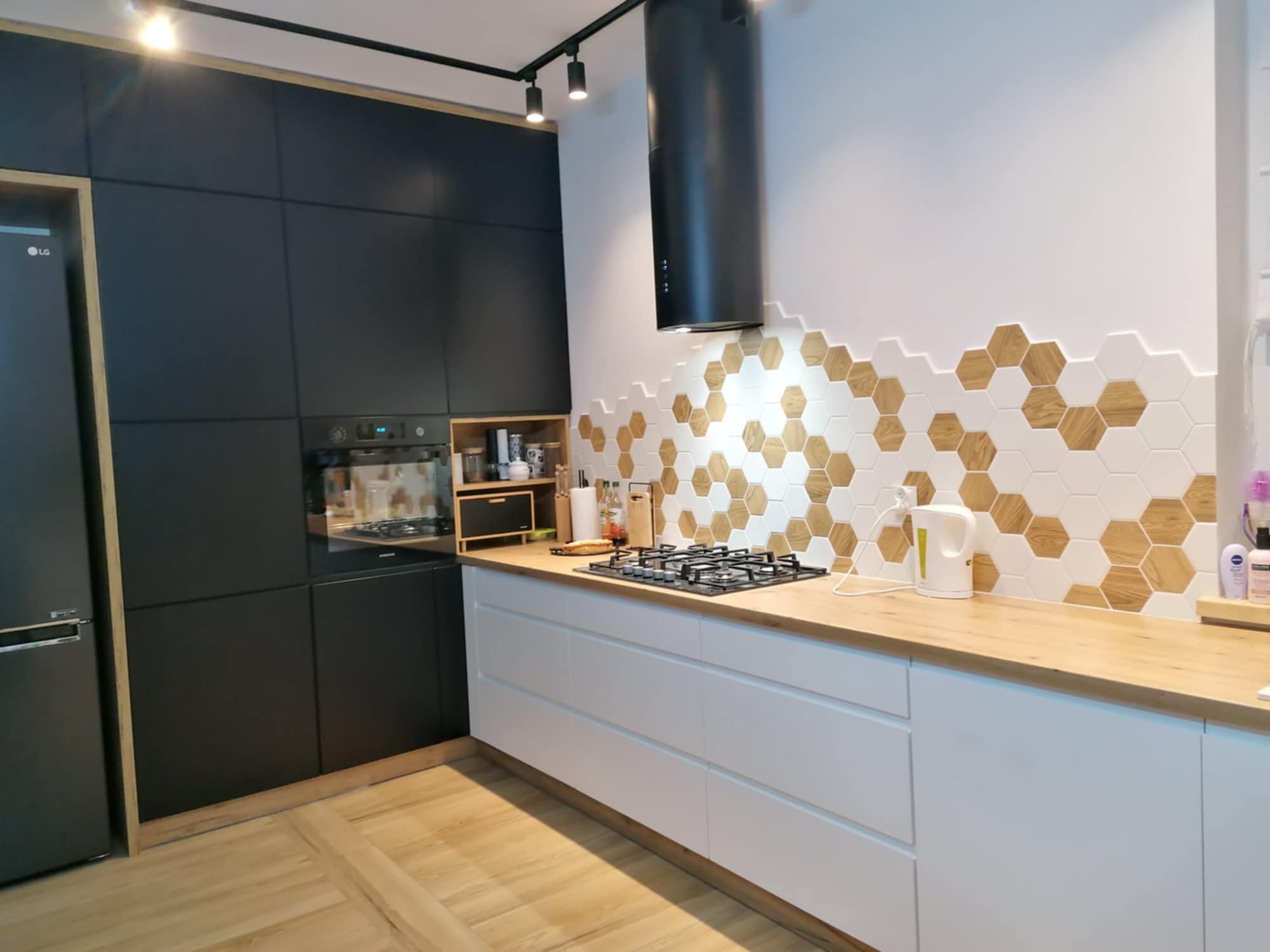 Nowoczesna kuchnia z czarnymi szafkami, drewnianym blatem i heksagonalnymi płytkami na ścianie, oświetlenie punktowe na suficie.
