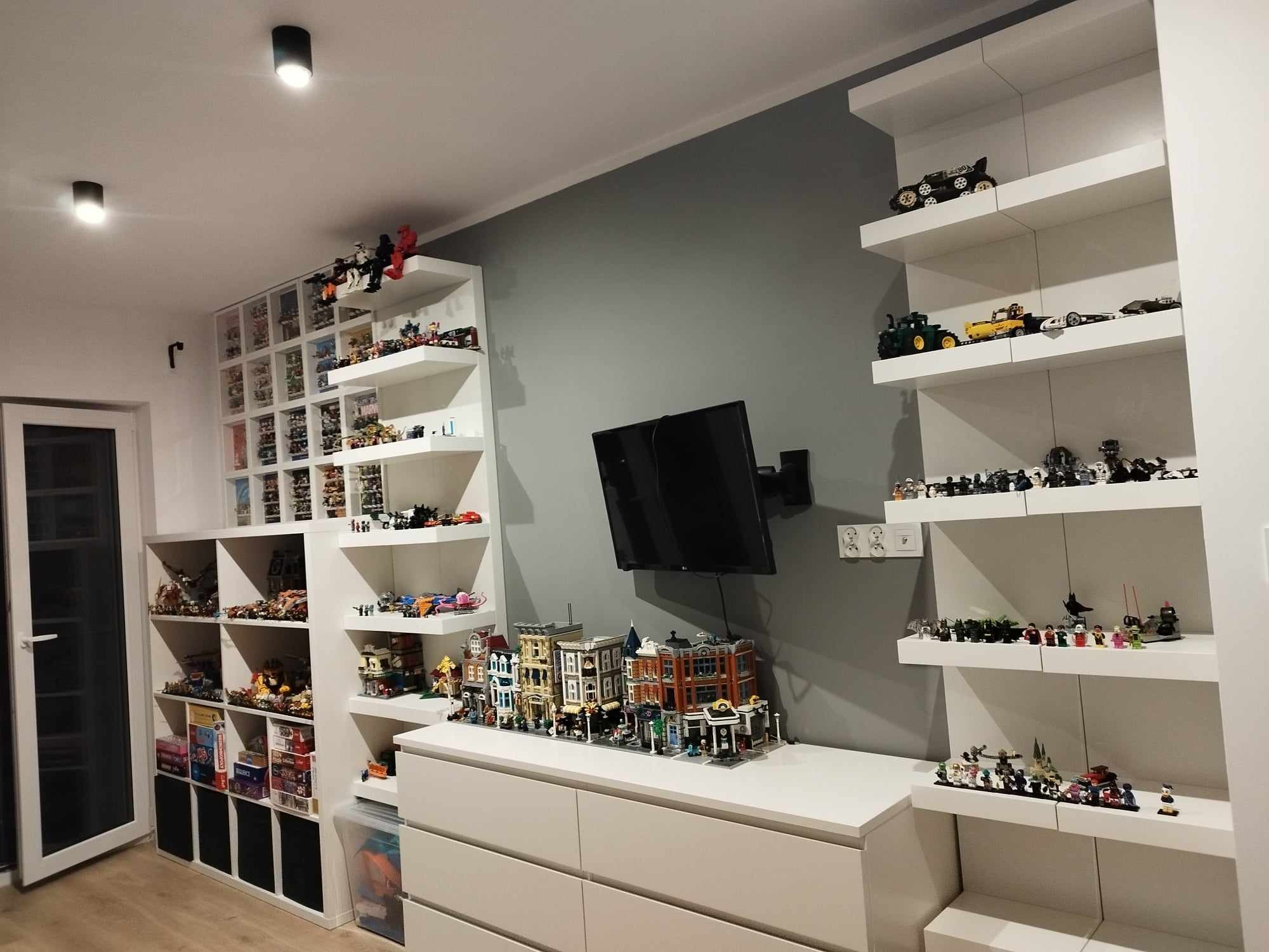 Pokój z białymi meblami i półkami, na których eksponowane są kolekcje klocków LEGO, w tym modele budynków i pojazdów, podwieszony telewizor na szarej ścianie.