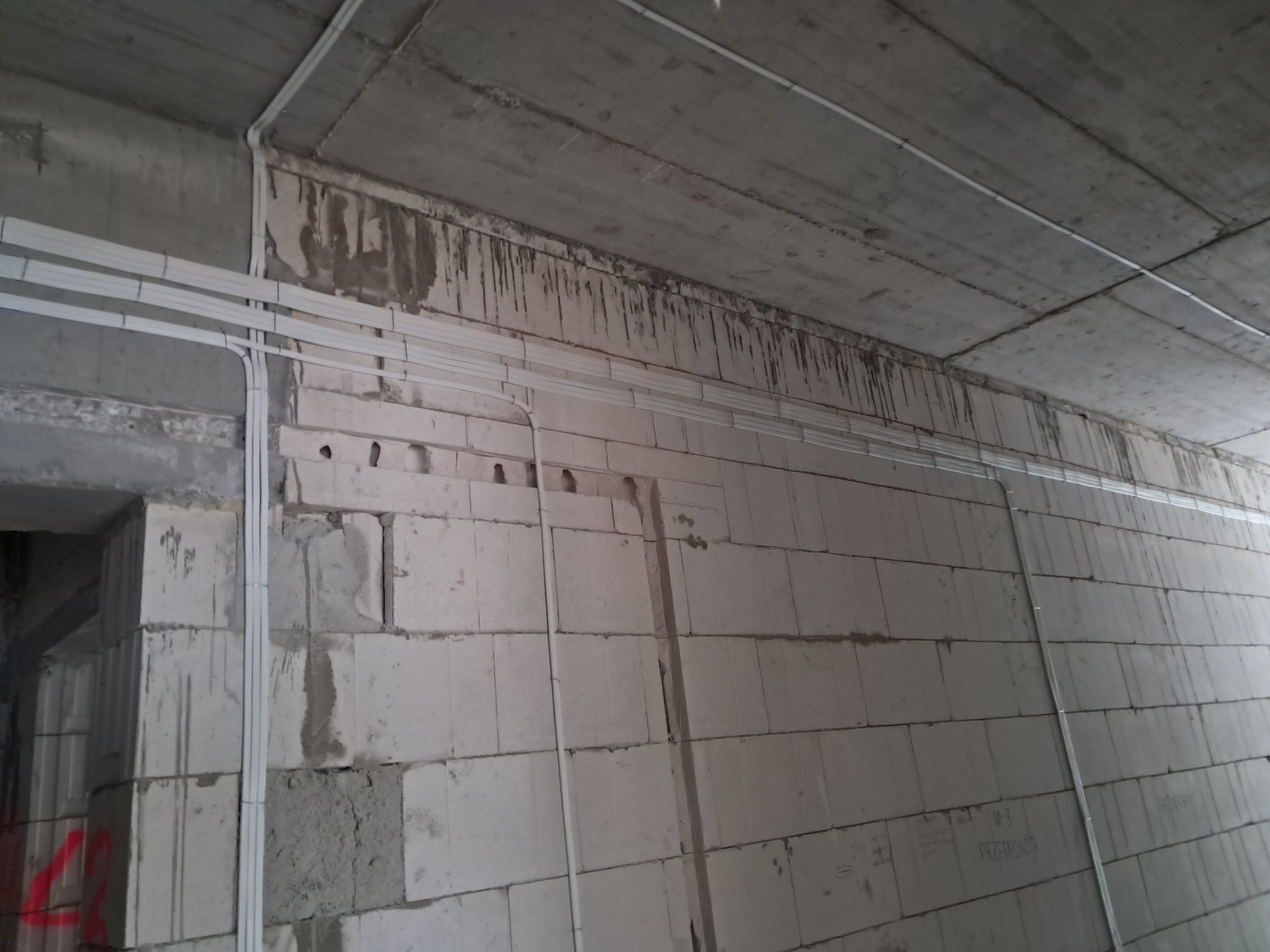 Surowy betonowy sufit i ściany z białych bloczków z widocznymi przewodami elektrycznymi w białych listwach, fragment otworu drzwiowego po lewej stronie.