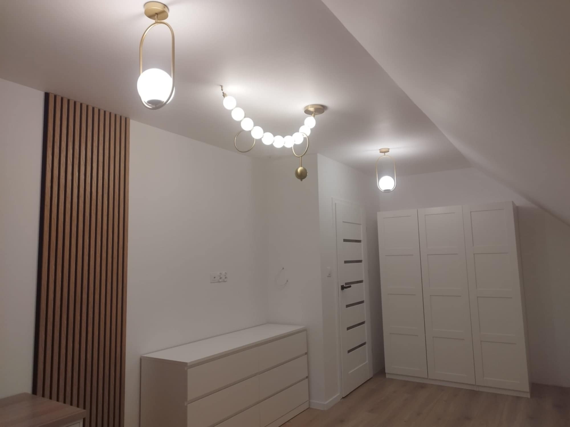 Pokój na poddaszu z drewnianą ścianką działową, białą komodą, szafą i designerskim oświetleniem.