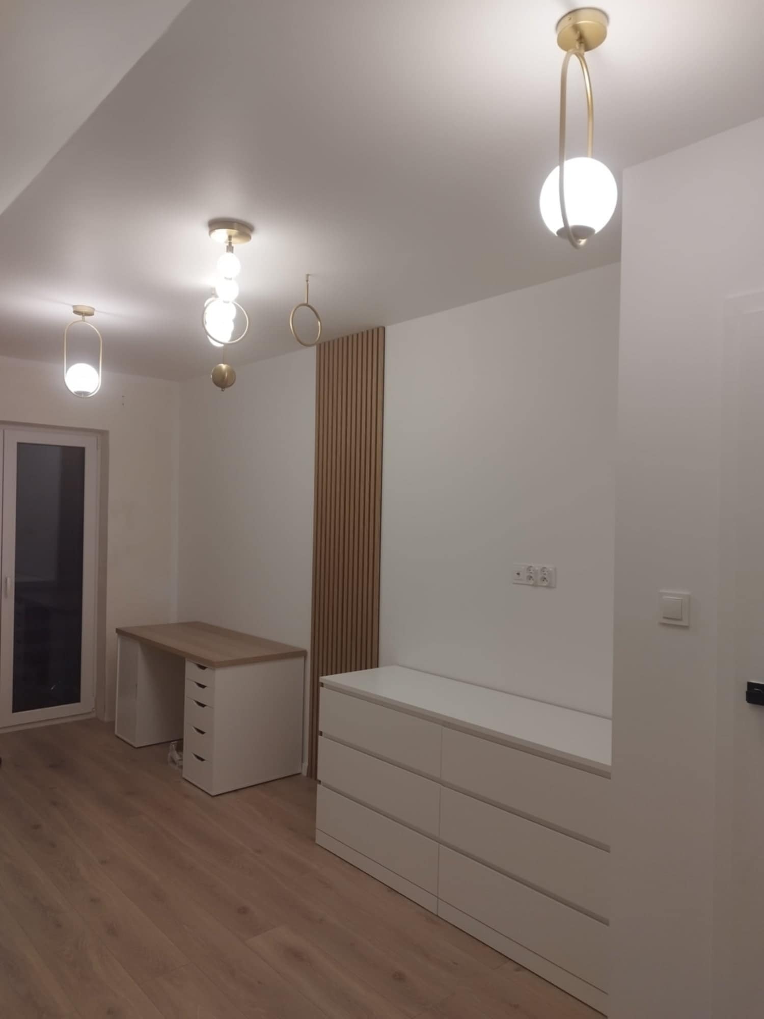 Pokój po remoncie: drewniana podłoga, białe ściany, biurko z szufladami, komoda oraz dekoracyjna drewniana listwa na ścianie i nowoczesne lampy sufitowe.