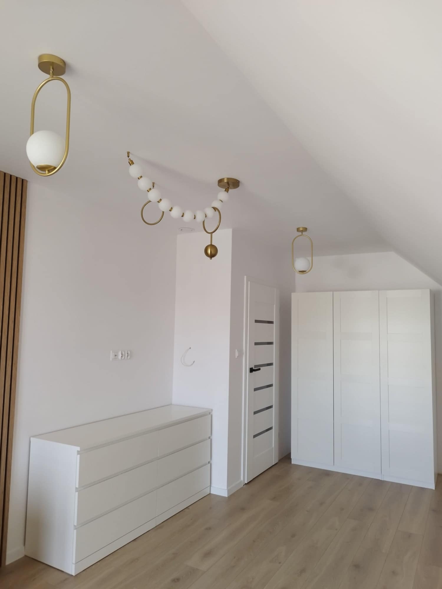 Jasne poddasze po remoncie z białymi meblami: komoda, szafa w zabudowie, drzwi z czarnymi paskami i trzema złotymi lampami o różnych kształtach.