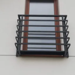 Czarna, metalowa balustrada okienna z poziomymi szczeblami, zamontowana na jasnej elewacji budynku przy brązowym oknie.