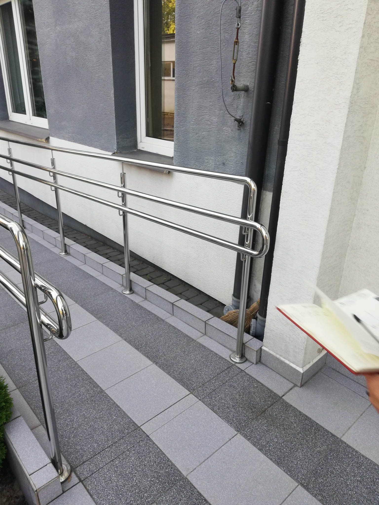 Zewnętrzna pochylnia dla wózków inwalidzkich z podwójną, chromowaną poręczą, widok z boku, z widocznym fragmentem notatnika w rogu kadru.