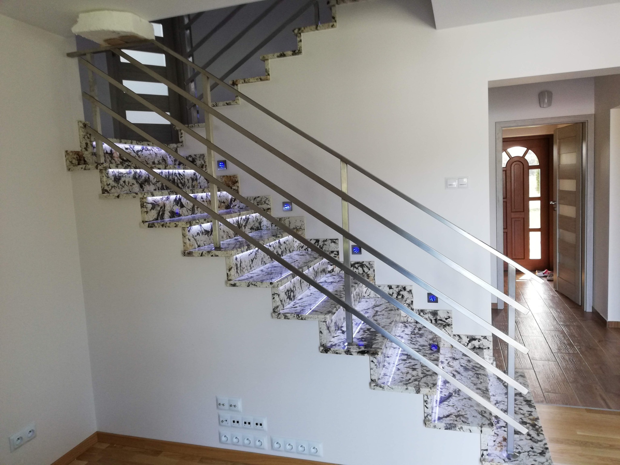 Nowoczesne schody z marmurowymi stopniami podświetlonymi niebieskim światłem LED, z metalową balustradą i widocznymi gniazdkami elektrycznymi na ścianie.