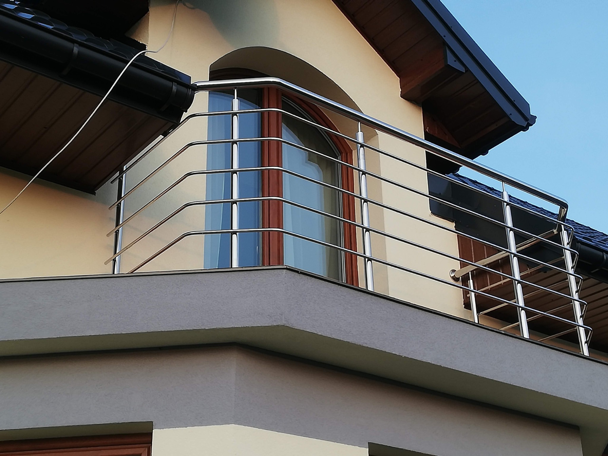 Balkon z nowoczesną, poziomą balustradą ze stali nierdzewnej, widok z dołu, na tle jasnej elewacji budynku z oknem o łukowatym kształcie.