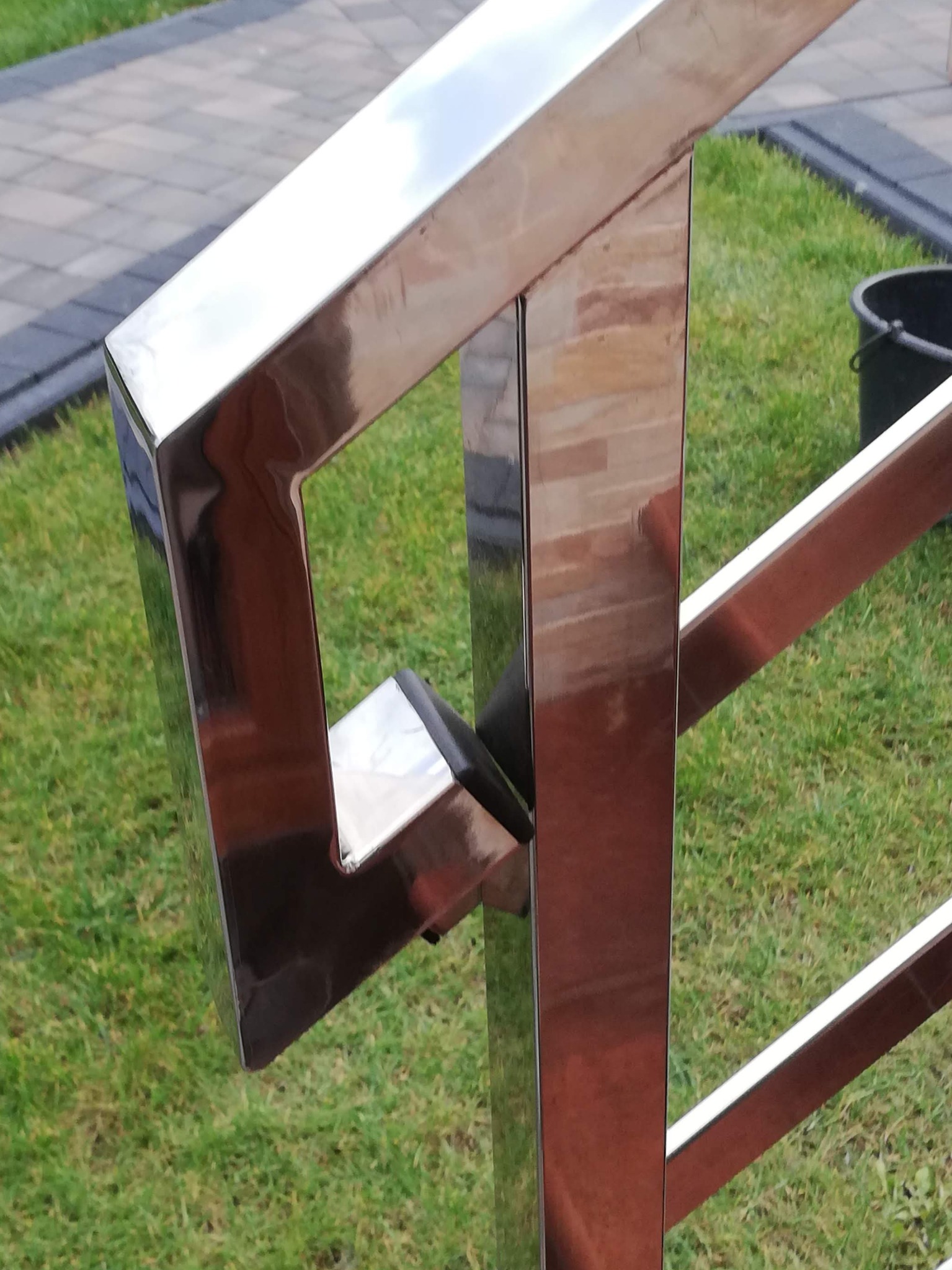 Detal narożnika balustrady nierdzewnej z widocznym mocowaniem do słupka; w tle trawnik i kostka brukowa.