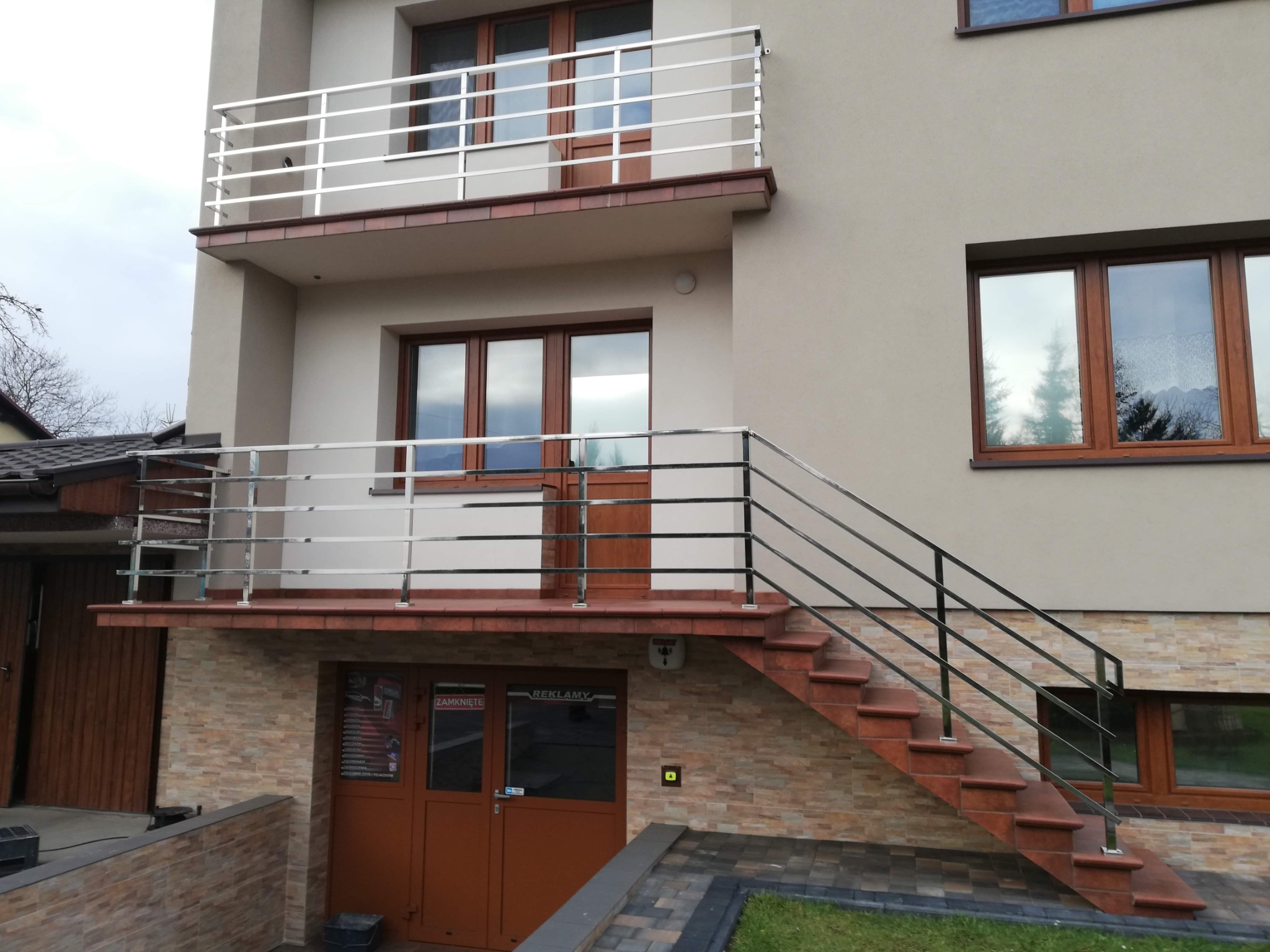 Dwukondygnacyjny budynek z balkonami zabezpieczonymi poziomymi, metalowymi balustradami oraz zewnętrznymi schodami z poręczą, prowadzącymi do wejścia na piętrze.