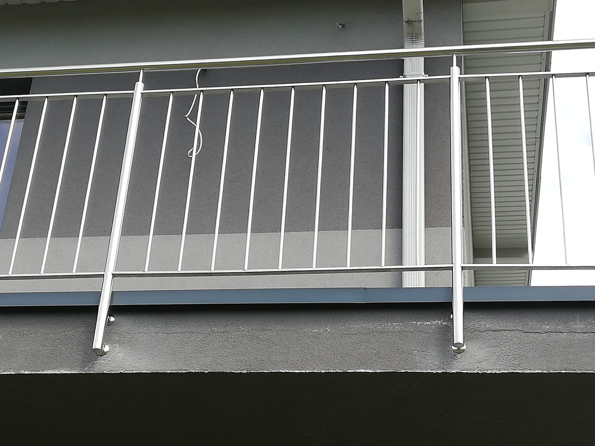 Balkon z nowoczesną, minimalistyczną balustradą ze stali nierdzewnej, widok z dołu, szary tynk strukturalny budynku w tle.