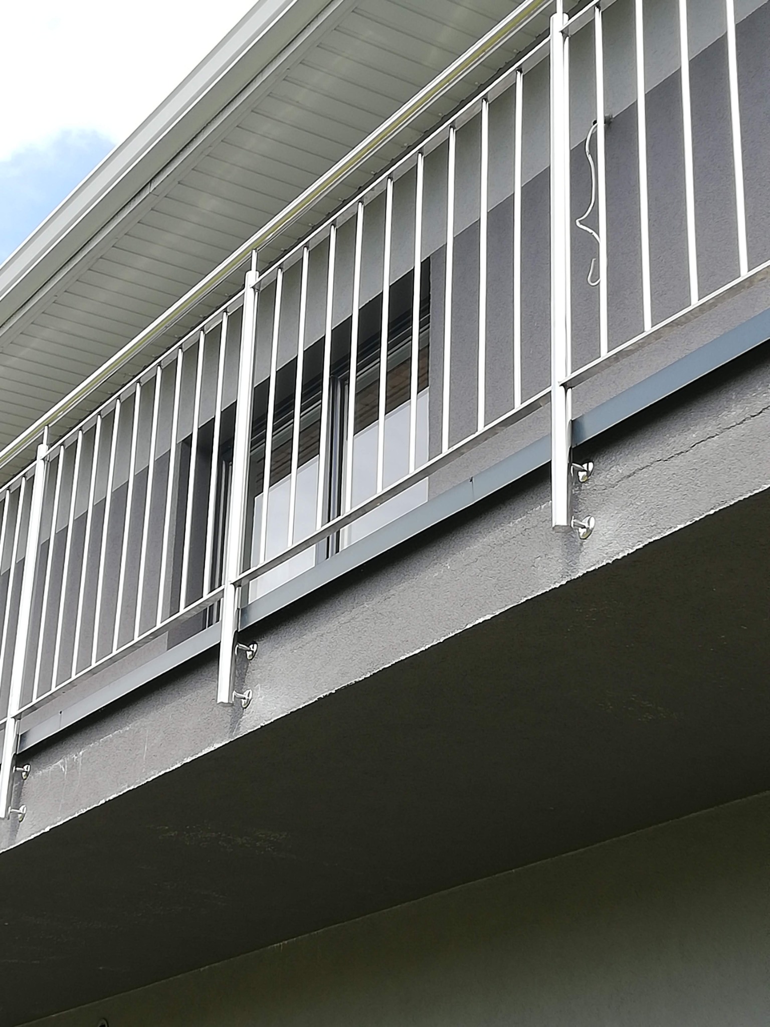 Balkon z nowoczesną, minimalistyczną balustradą ze stali, widok z dołu, z widocznymi mocowaniami do betonowej konstrukcji balkonu, na jednej z pionowych rurek wisi haczyk.