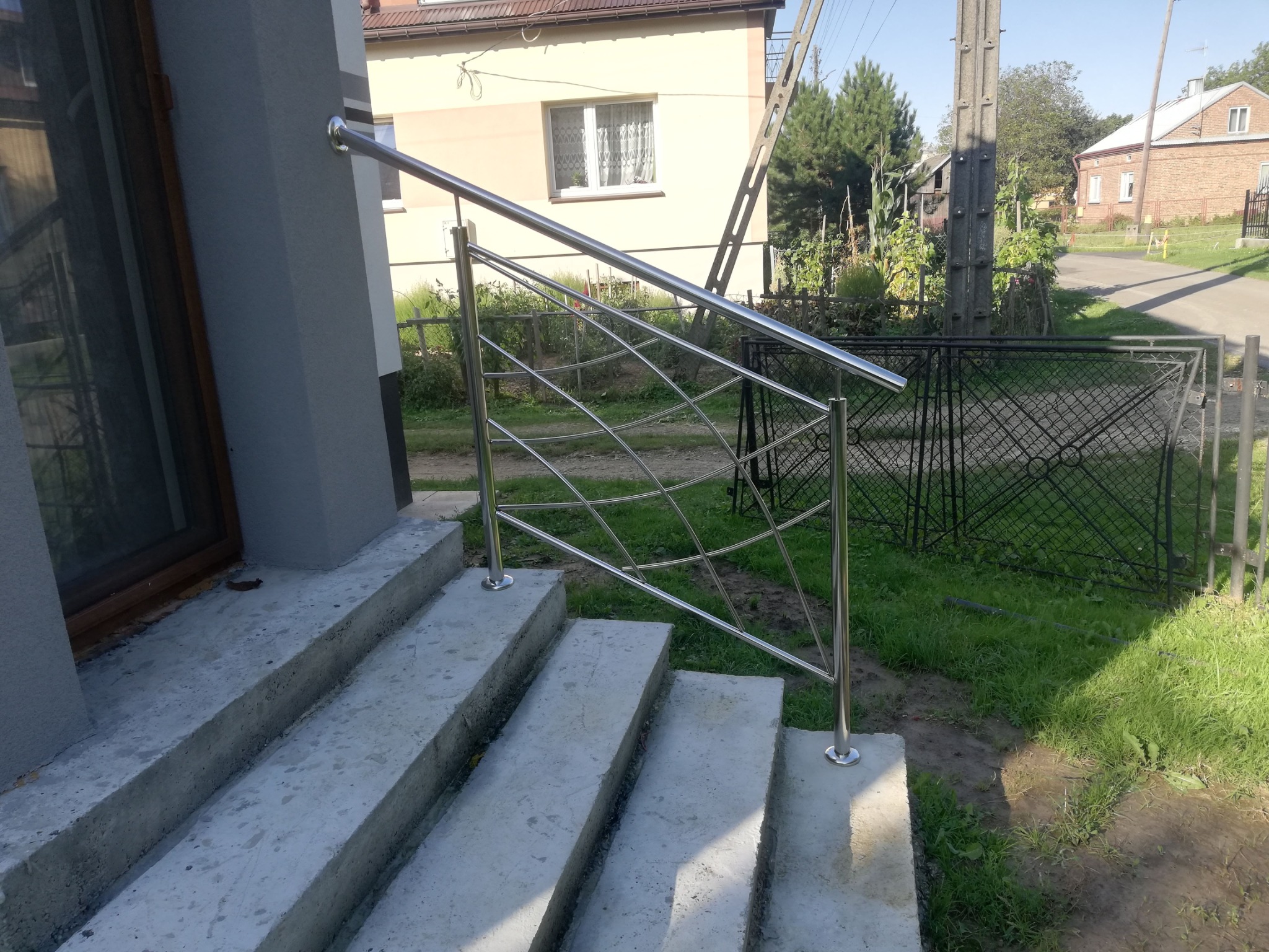 Polerowana, metalowa poręcz schodowa z nietypowym, krzyżowym wypełnieniem, zamontowana przy betonowych schodach wejściowych do domu.