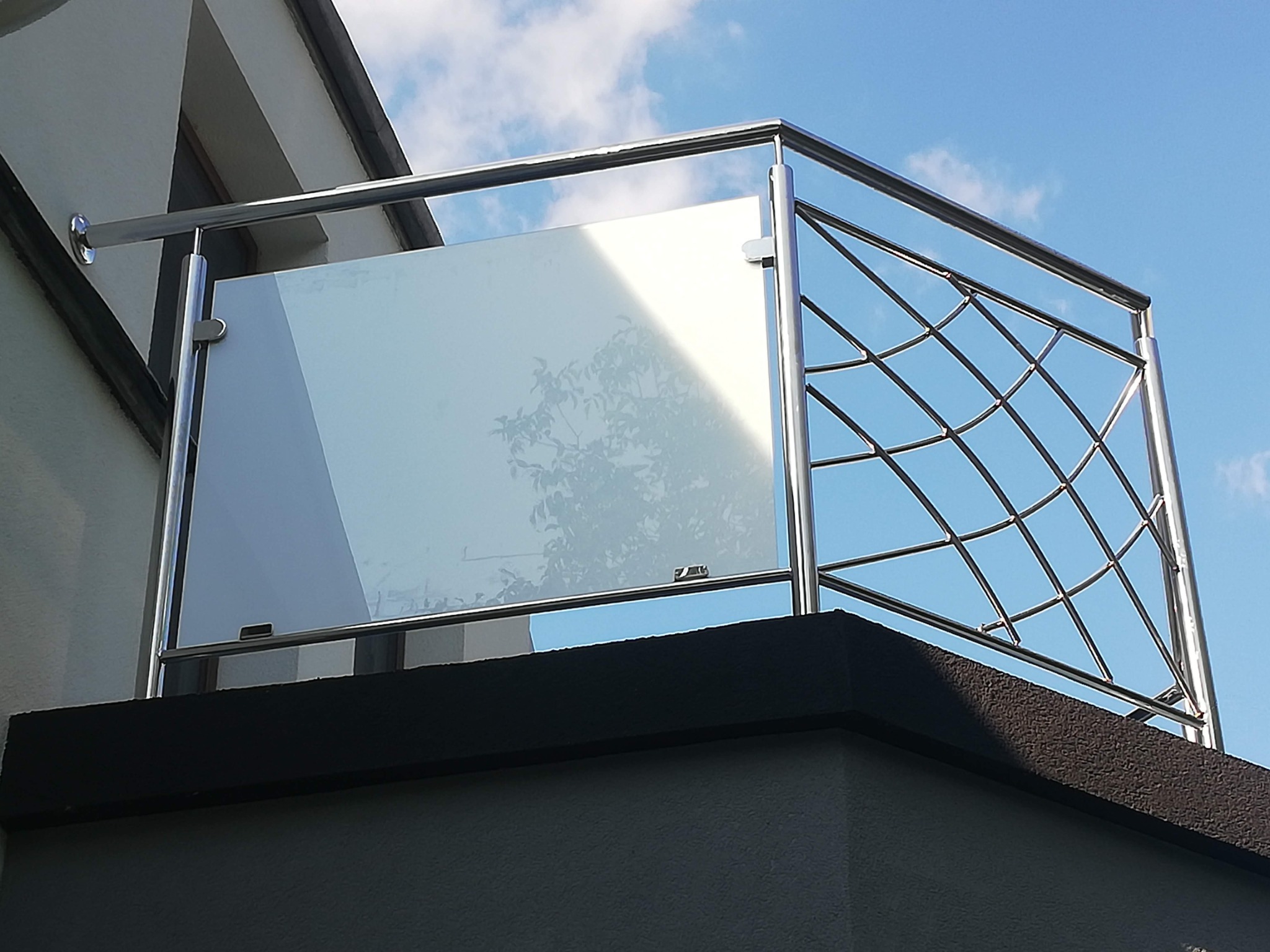 Balkon z nowoczesną balustradą ze stali nierdzewnej z wypełnieniem szklanym i ażurowym metalowym, widok z dołu na tle błękitnego nieba.