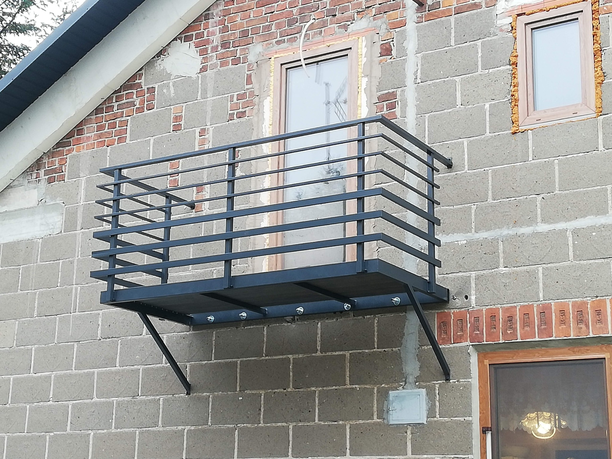 Czarny, metalowy balkon z prostą balustradą przymocowany do ściany budynku z cegły i pustaków, widok z zewnątrz na elewację.