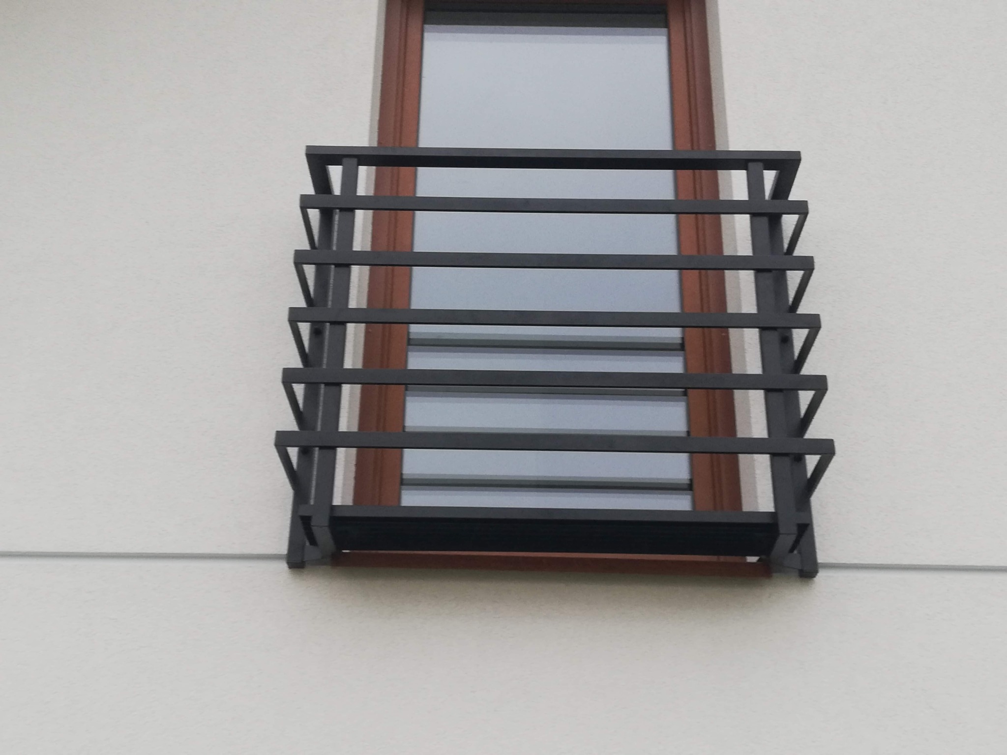 Czarna, metalowa balustrada okienna z poziomymi szczeblami, zamontowana na jasnej elewacji budynku przy brązowym oknie.