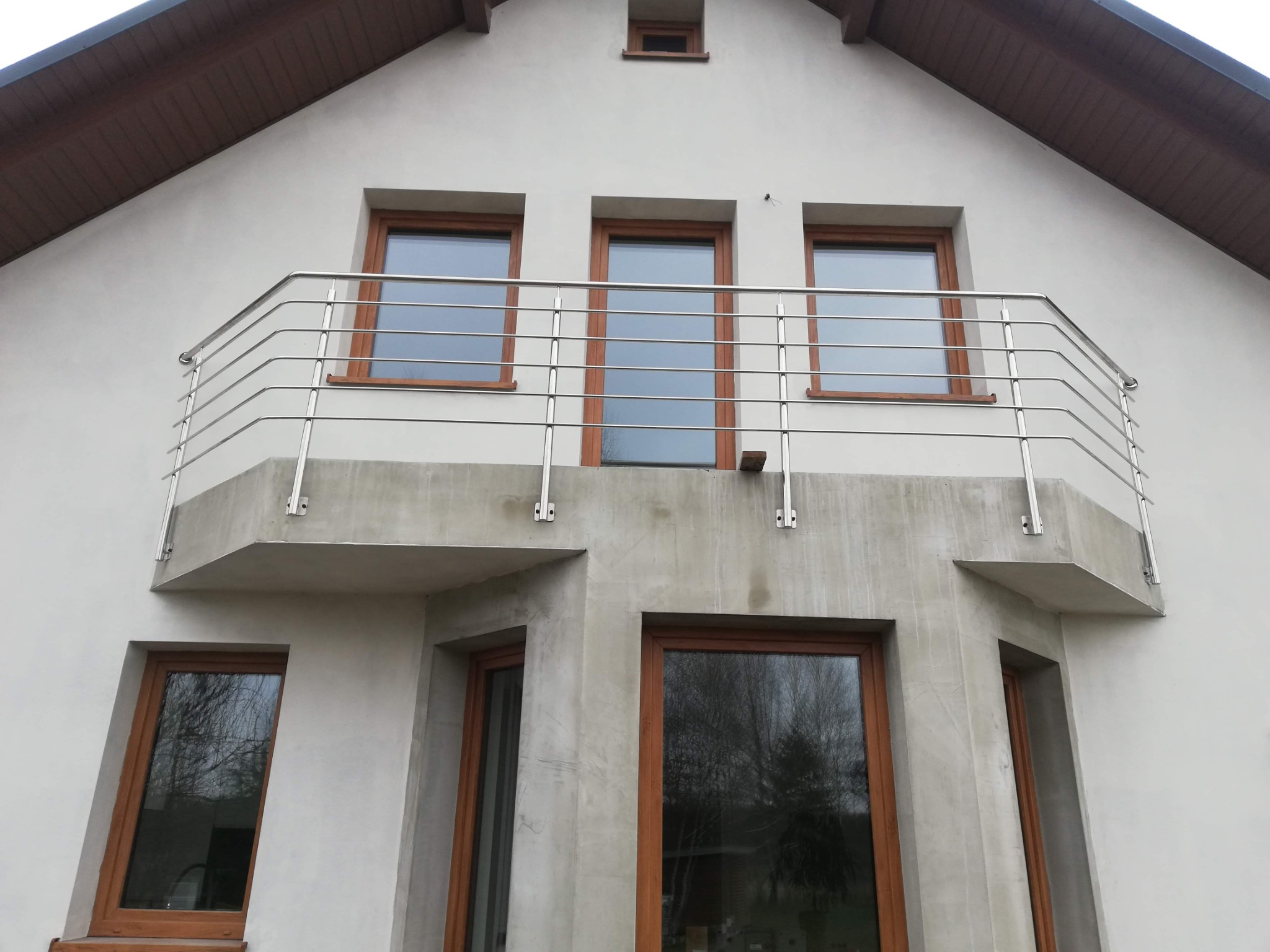 Balkon z prostą, metalową balustradą z rurek, zamontowaną na betonowej konstrukcji, widoczne trzy okna z brązowymi ramami na górze i dwa na dole, biała fasada budynku z brązowym dachem.