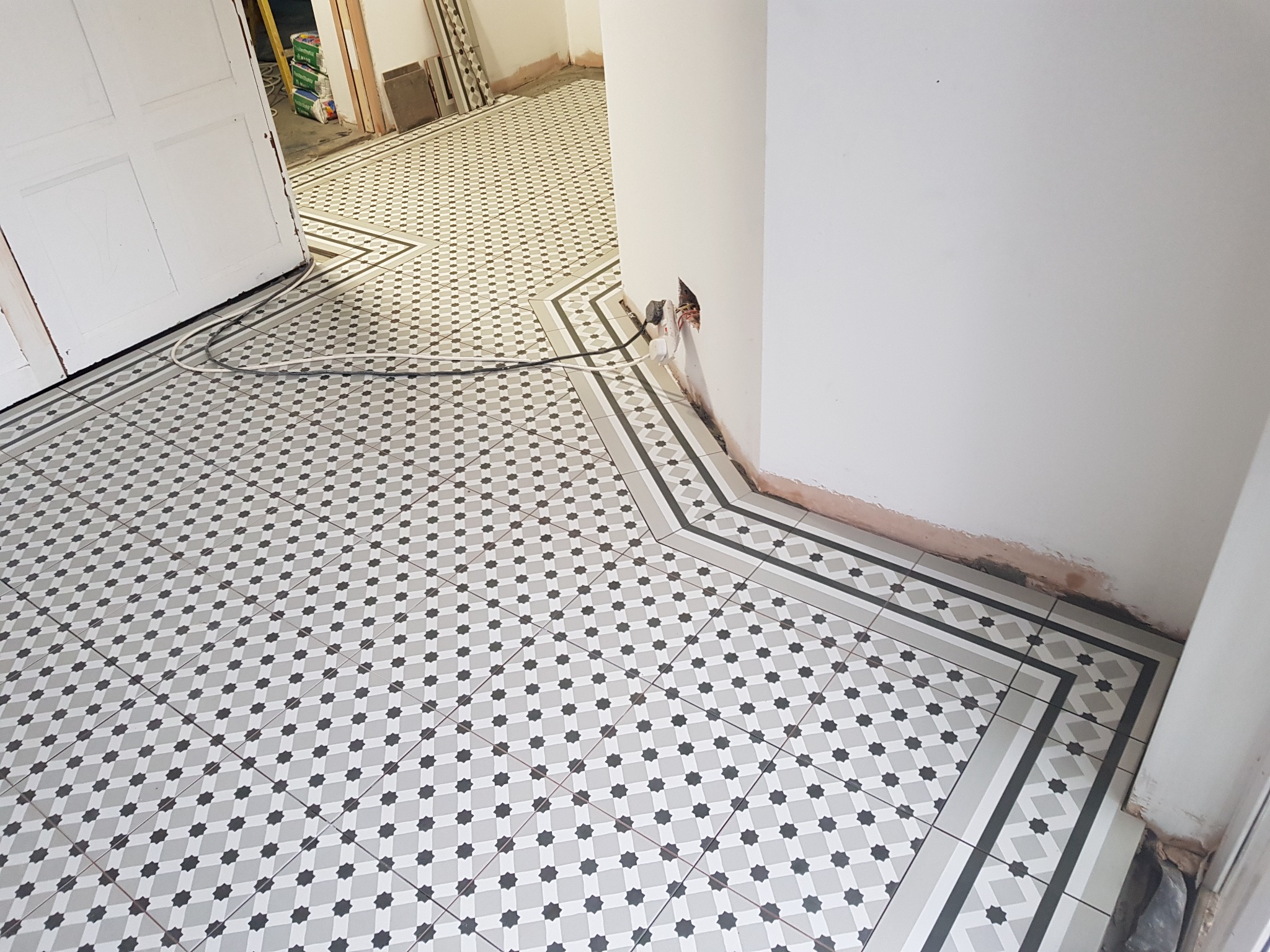 Ułożona podłoga z płytek w biało-szare wzory. Fragment ściany z wystającą instalacją elektryczną. Widoczne otwarte drzwi i dalsza część podłogi.