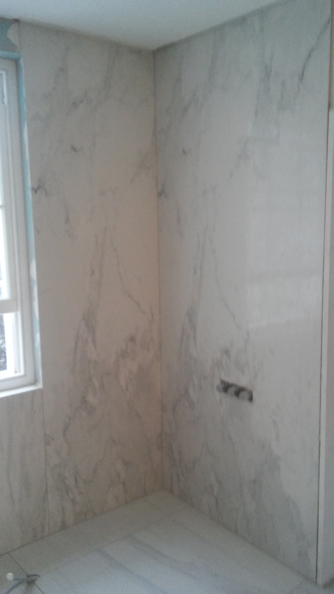 Wykończona łazienka z marmurowymi płytkami na ścianach i podłodze, widoczne okno i wystający ze ściany fragment instalacji.