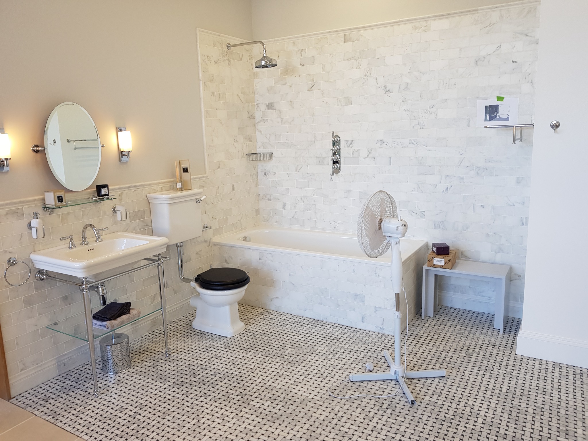 Elegancka łazienka w bieli i szarości: mozaika na podłodze, marmurowe płytki na ścianach, klasyczna umywalka na metalowym stelażu i wanna. Całość doświetlona lampami.