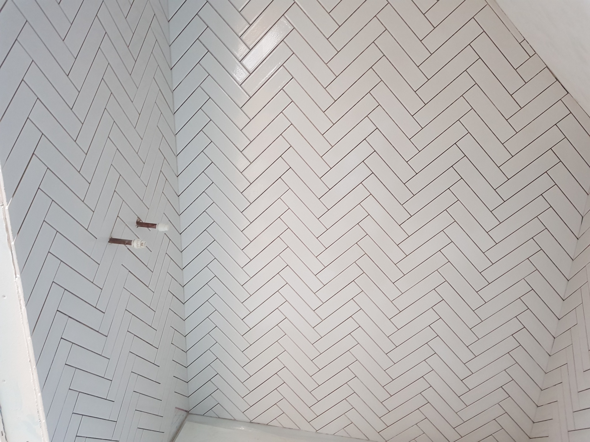 Białe, prostokątne płytki ułożone w jodełkę na ścianie łazienki. Widoczne wystające rurki instalacji. Jasne, równomierne oświetlenie.