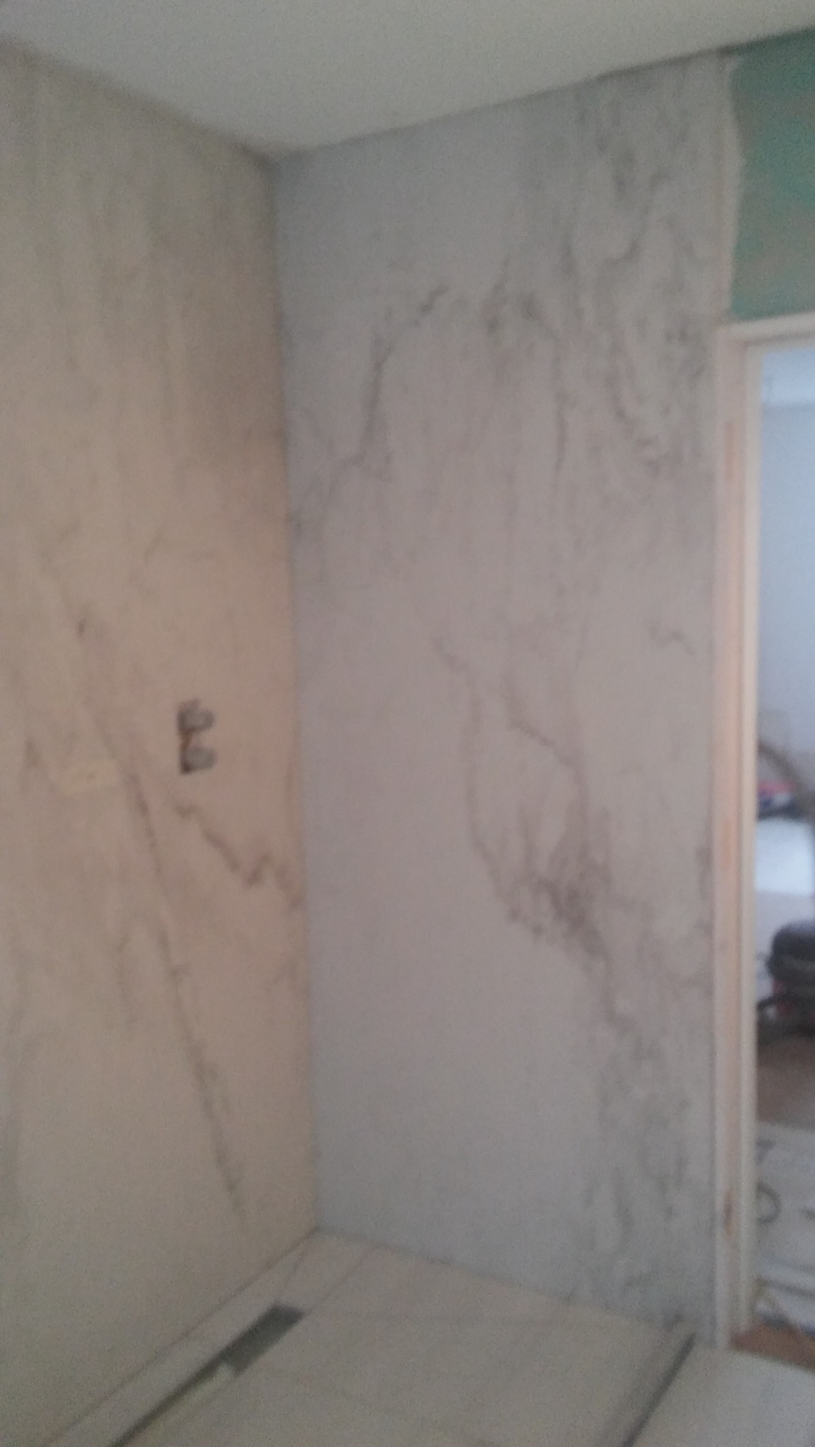 Wykończona łazienka z marmurowymi płytkami na ścianach i podłodze. Widoczny odpływ liniowy i fragment drzwi do innego pomieszczenia.