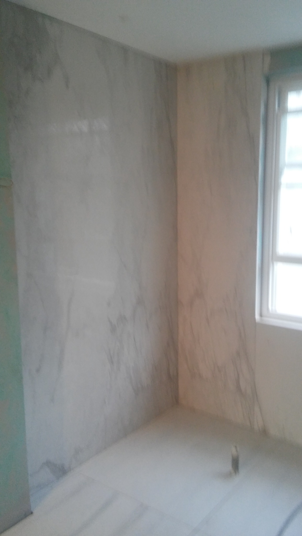 Wykończona łazienka z marmurowymi płytkami na ścianach i podłodze, okno po prawej stronie. Neutralna kolorystyka, minimalistyczny design. Widok na narożnik pomieszczenia.