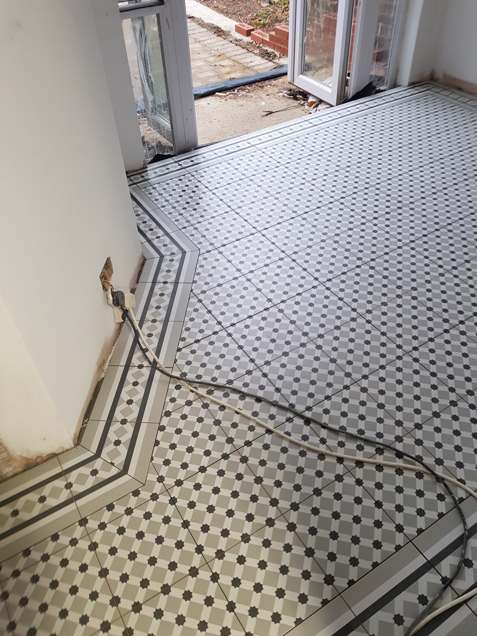 Ułożona podłoga z płytek w geometryczny wzór gwiazd, z widocznym fragmentem ściany z gniazdkiem i kablami oraz otwartymi drzwiami balkonowymi.