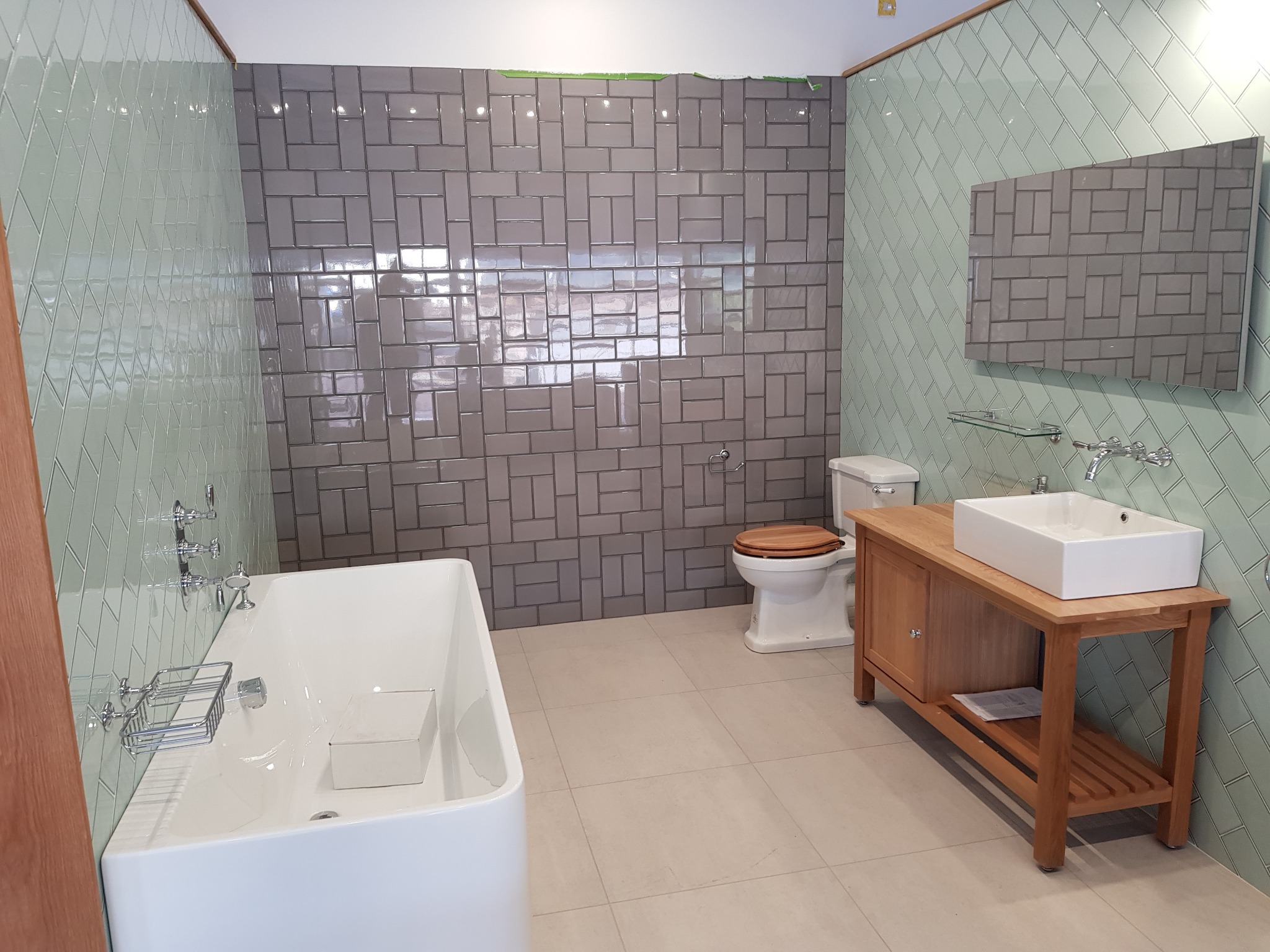 Wykończona łazienka z wanną, umywalką na drewnianej szafce i toaletą. Ściany wyłożone płytkami w odcieniach szarości i zieleni ułożonymi we wzory.