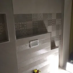 Fragment ściany łazienki z ułożonymi płytkami w dw&oacute;ch stylach: gładkie duże płyty oraz dekoracyjne mozaiki u g&oacute;ry, widoczne rury instalacji wodnej.