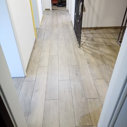 Łukasz Turek Usługi ogólnobudowlane - Korytarz z nową, jasną podłogą z płytek imitujących drewno, widoczna odkurzacz przemysłowy w tle i zabezpieczone żółtą taśmą malarską ściany.