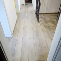 Korytarz z nową, jasną podłogą z płytek imitujących drewno, widoczna odkurzacz przemysłowy w tle i zabezpieczone żółtą taśmą malarską ściany.